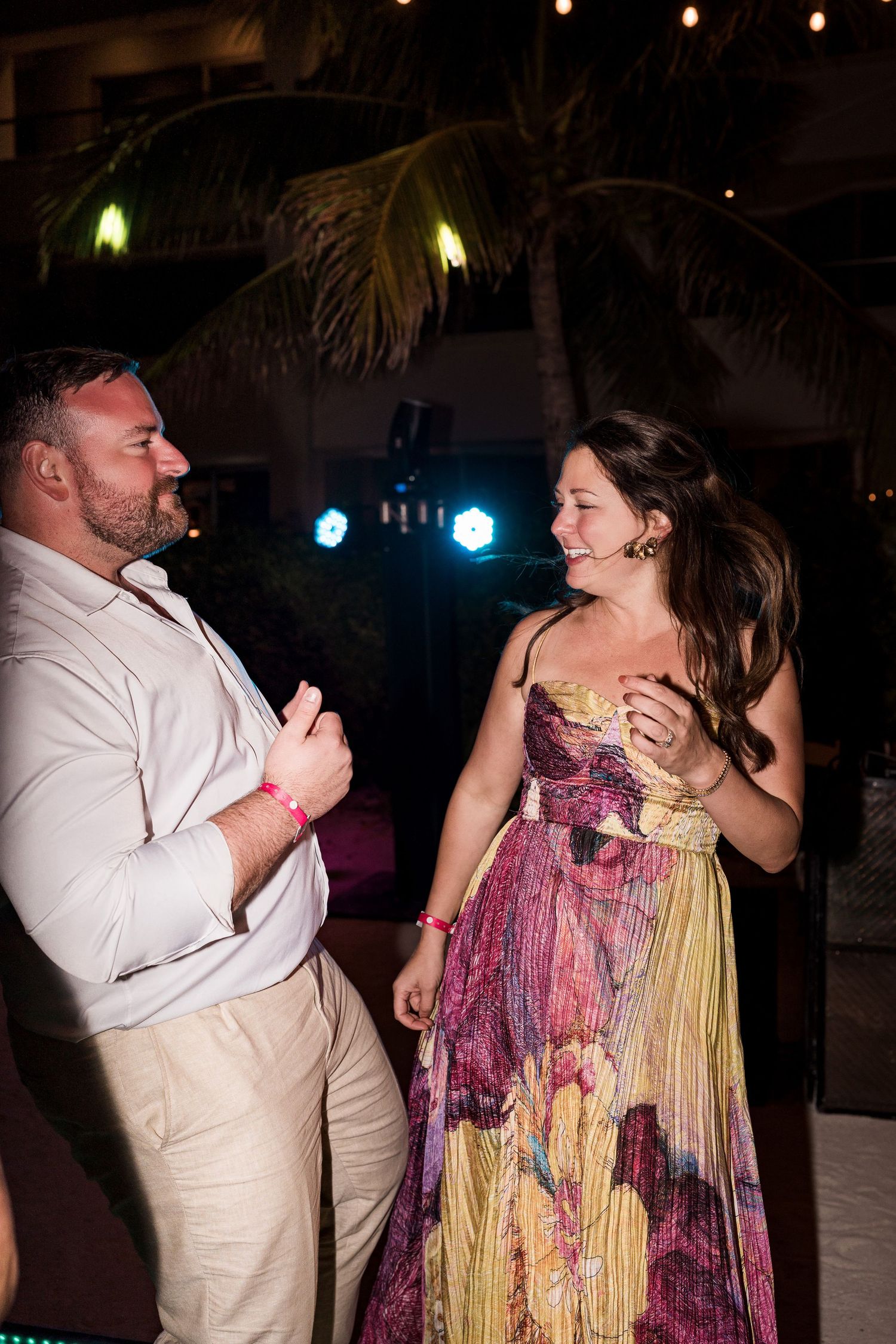 Couple dancing and laughing together at an outdoor evening wedding reception with palm trees and colorful lights.
