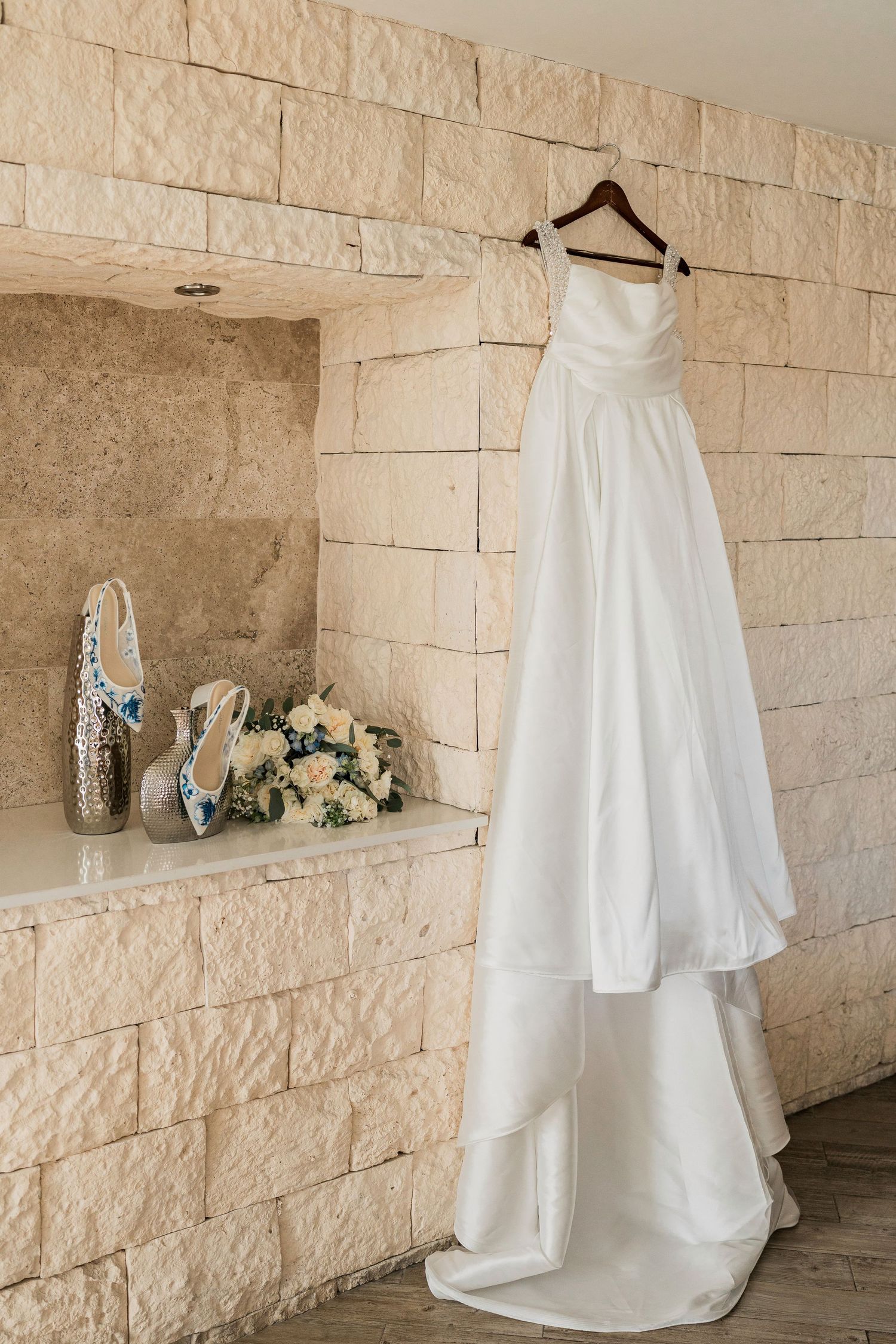 White wedding dress hanging on wooden hanger against stone wall, with bridal shoes and bouquet displayed nearby.
