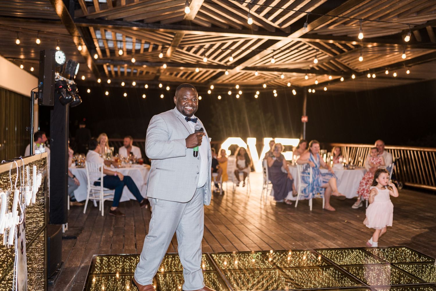 Man in gray suit giving speech at elegant wedding reception with string lights and LOVE sign backdrop.