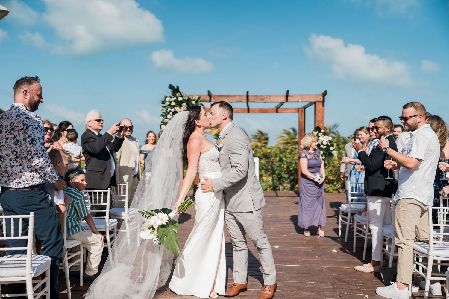 Bride and groom share first kiss at outdoor rooftop wedding ceremony with guests cheering under blue sky.