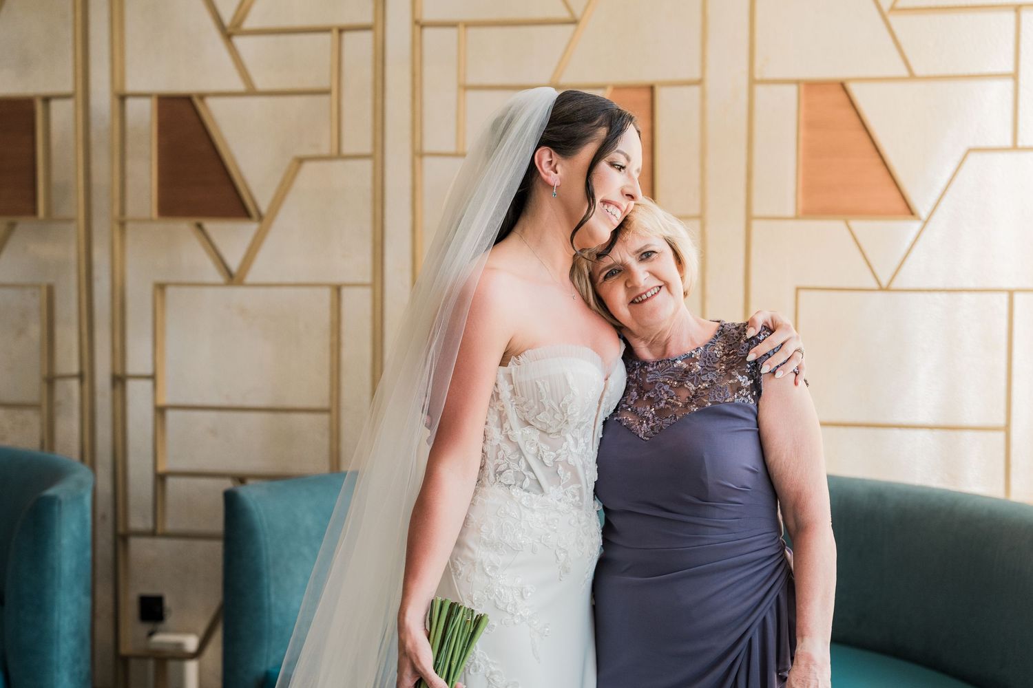 Bride in white lace gown and veil embraces older woman in purple dress, sharing emotional moment before wedding ceremony.