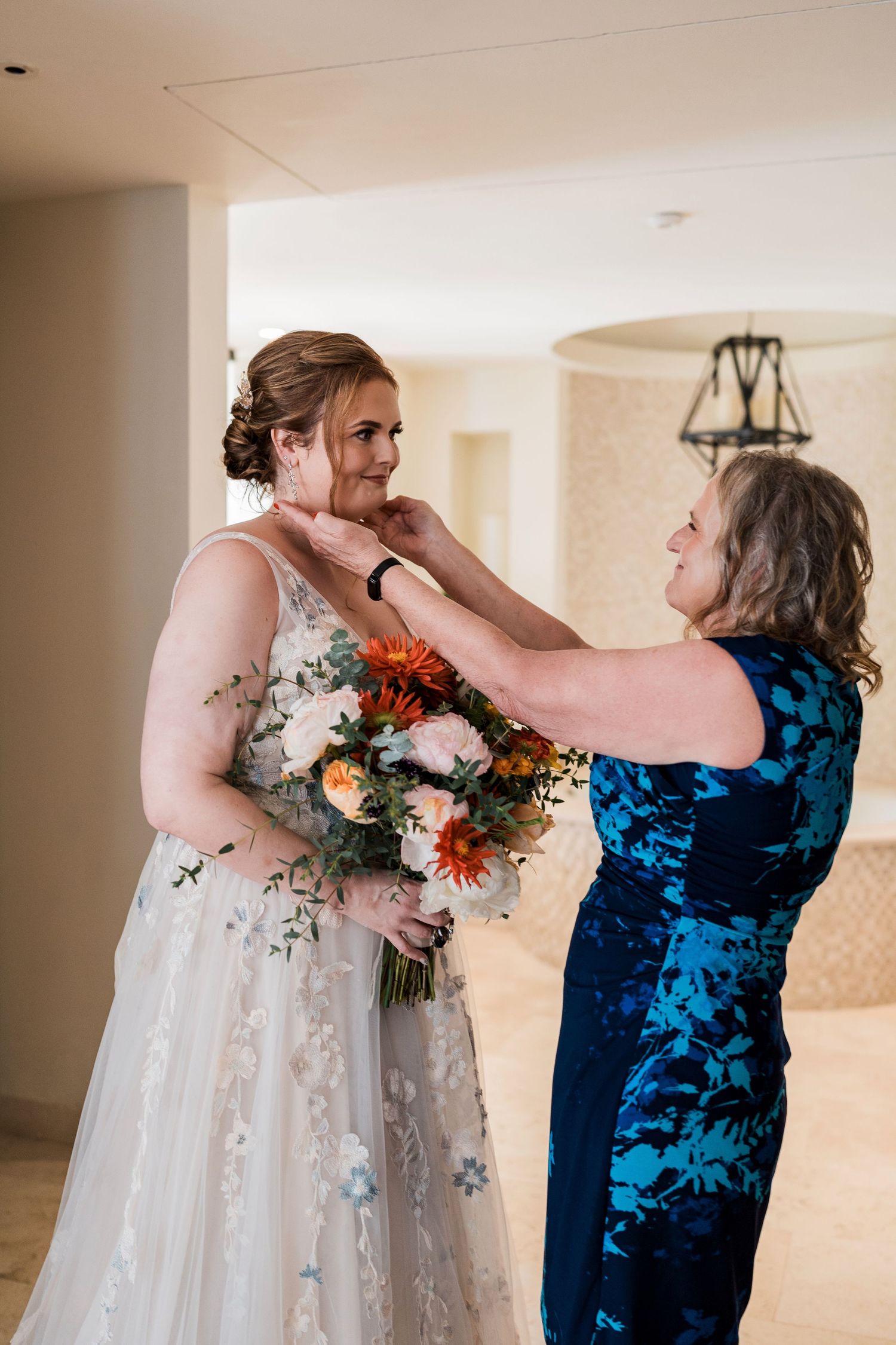 Bride in floral gown holds bouquet as woman in blue dress adjusts her necklace before wedding ceremony.