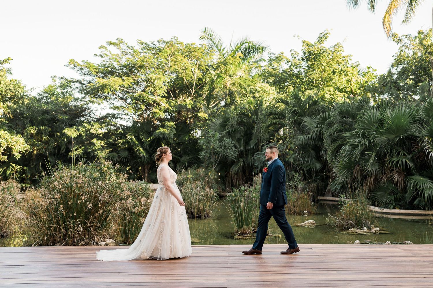 Secrets Akumal Wedding - Bride and groom walking on wooden deck surrounded by lush tropical greenery and pond at outdoor wedding venue.