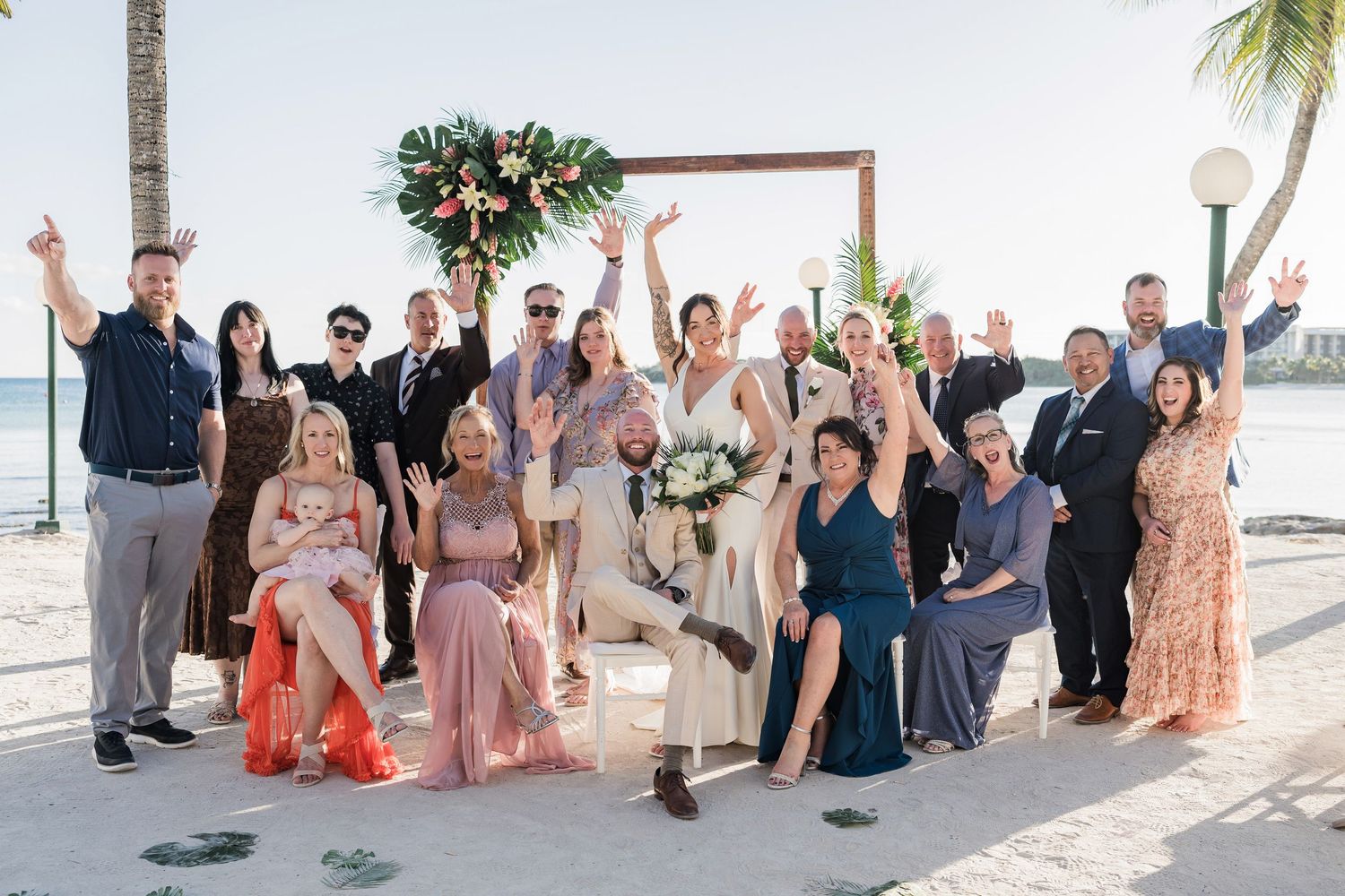 Wedding party celebrates on a tropical beach with a floral arch, guests cheering in colorful formal attire.
