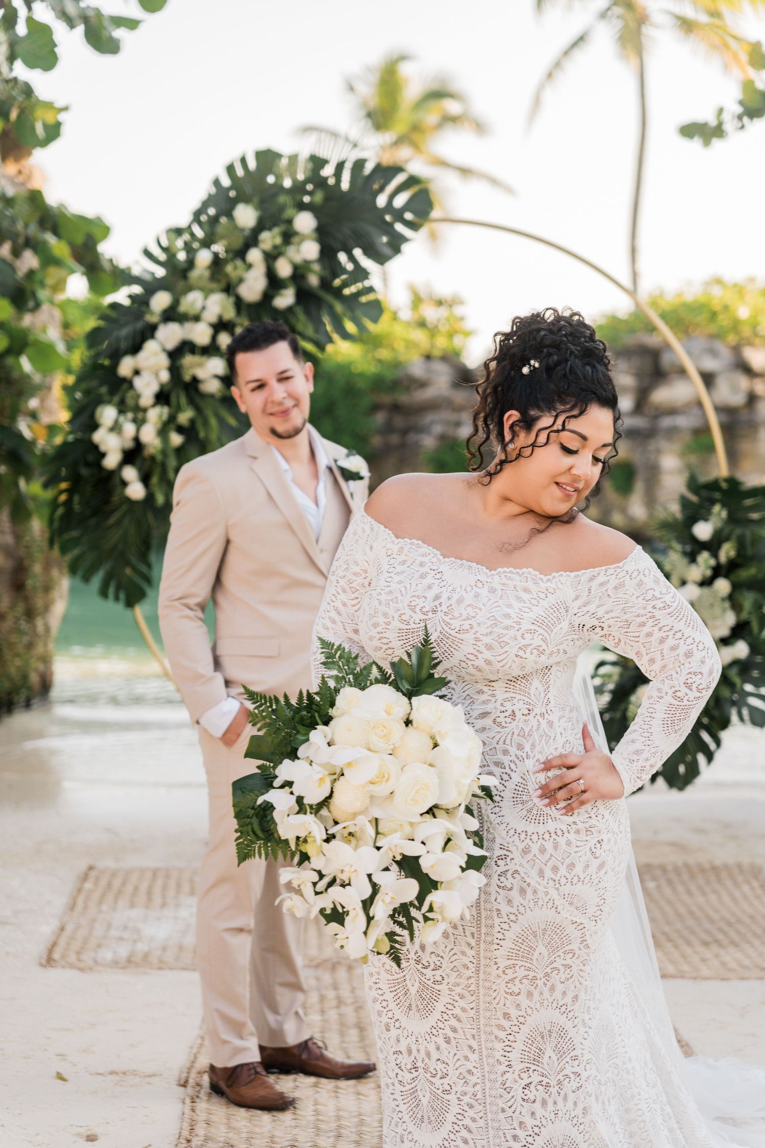 Xcaret Arte Wedding - Bride in lace off-shoulder gown holds white bouquet as groom in tan suit stands before floral arch on tropical beach.