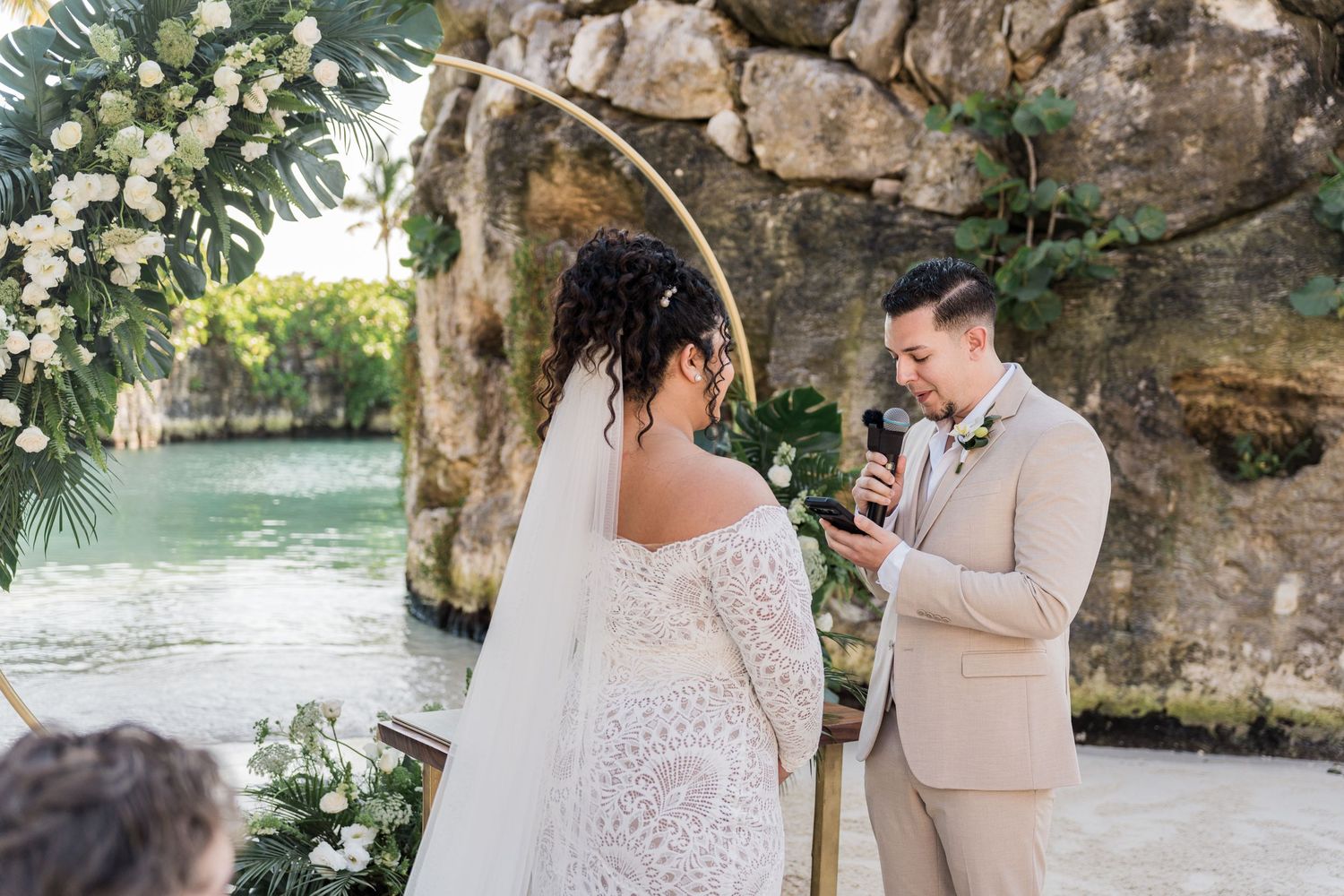 Xcaret Arte Wedding - Groom reads vows to bride in white lace dress at outdoor waterfront wedding ceremony with floral arch.