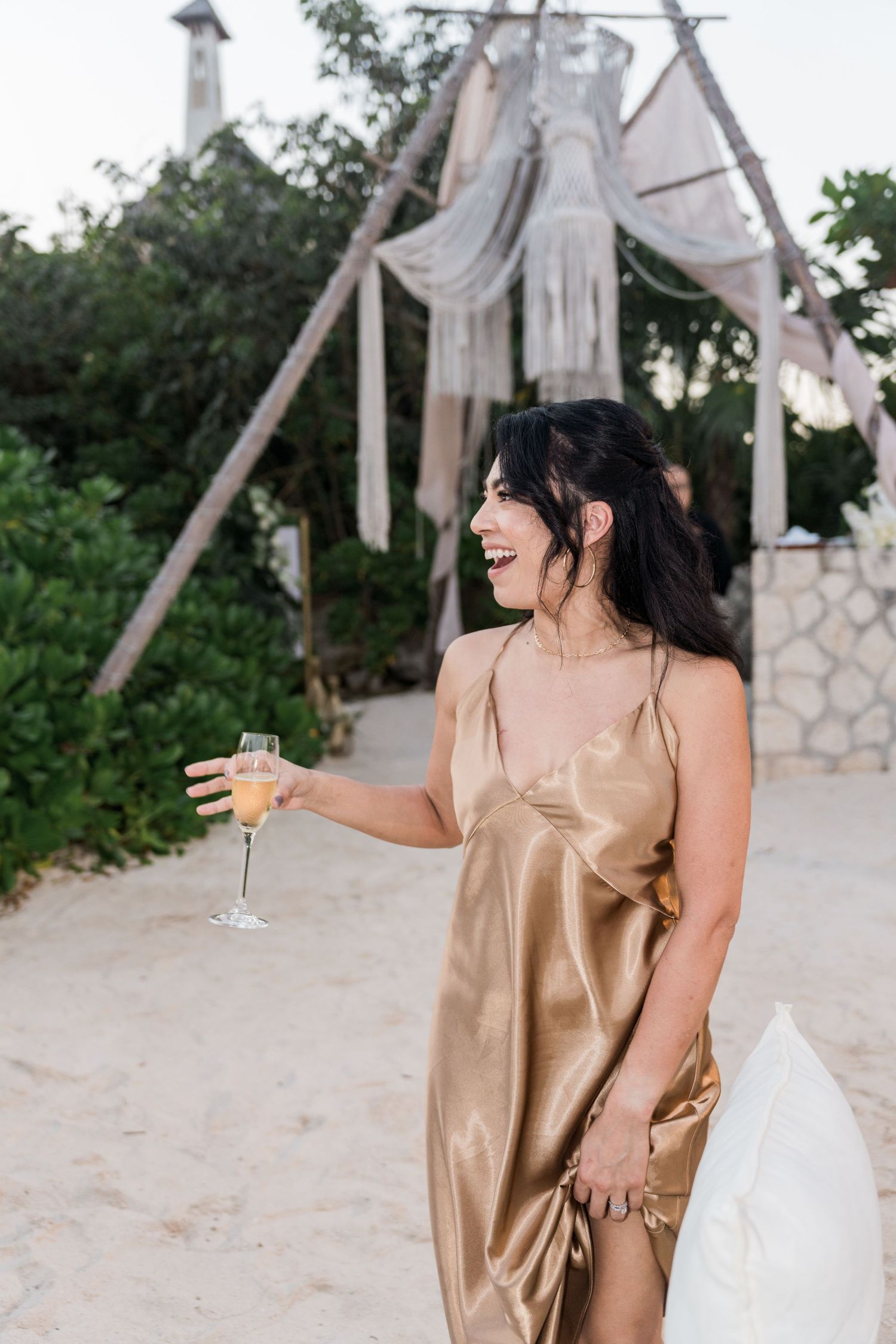 Xcaret Arte Wedding - Woman in gold satin dress holding champagne glass laughing at beach wedding celebration.
