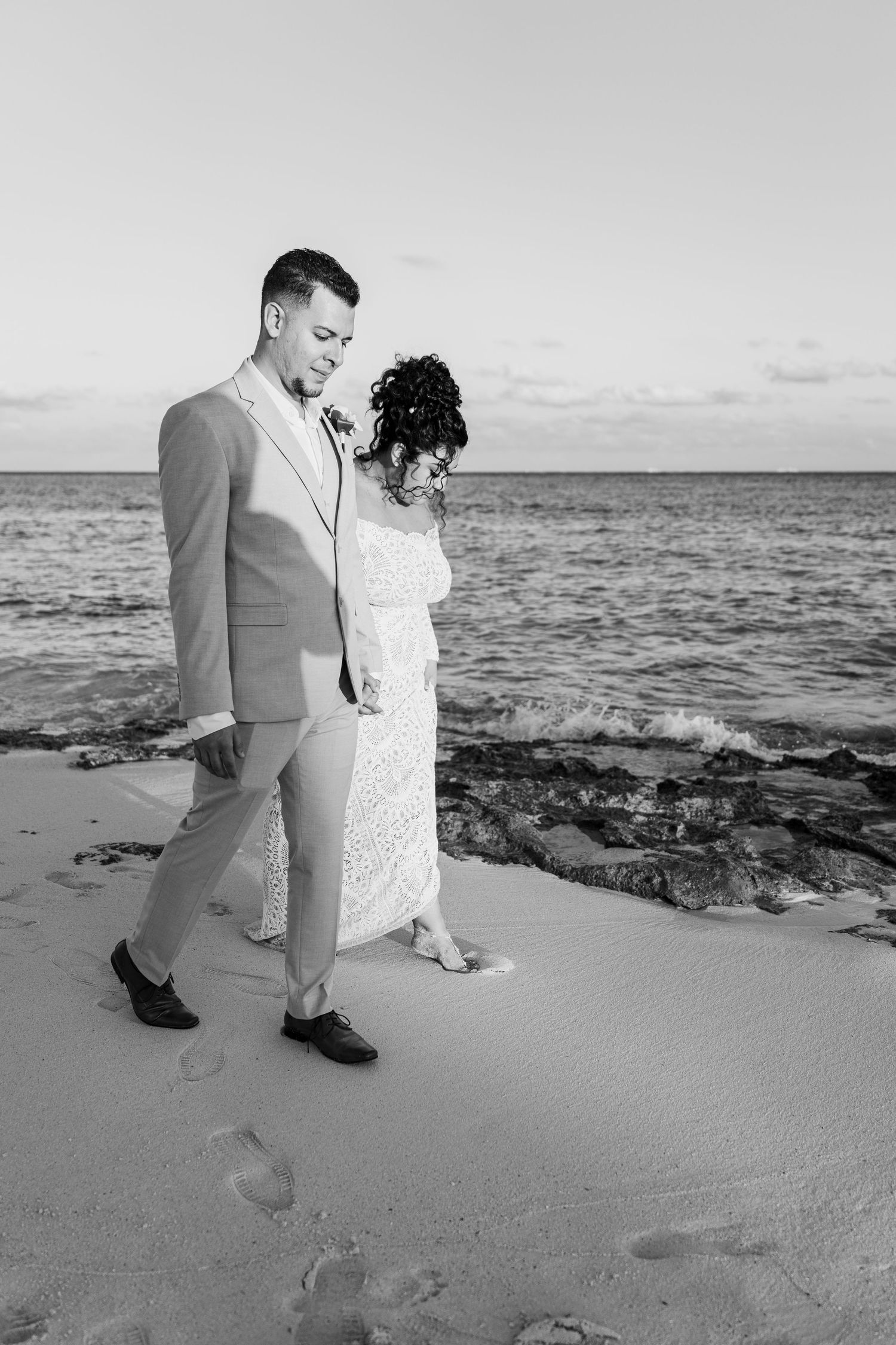 Xcaret Arte Wedding - Black and white photo of a bride and groom walking hand in hand on a sandy beach with ocean waves behind them.