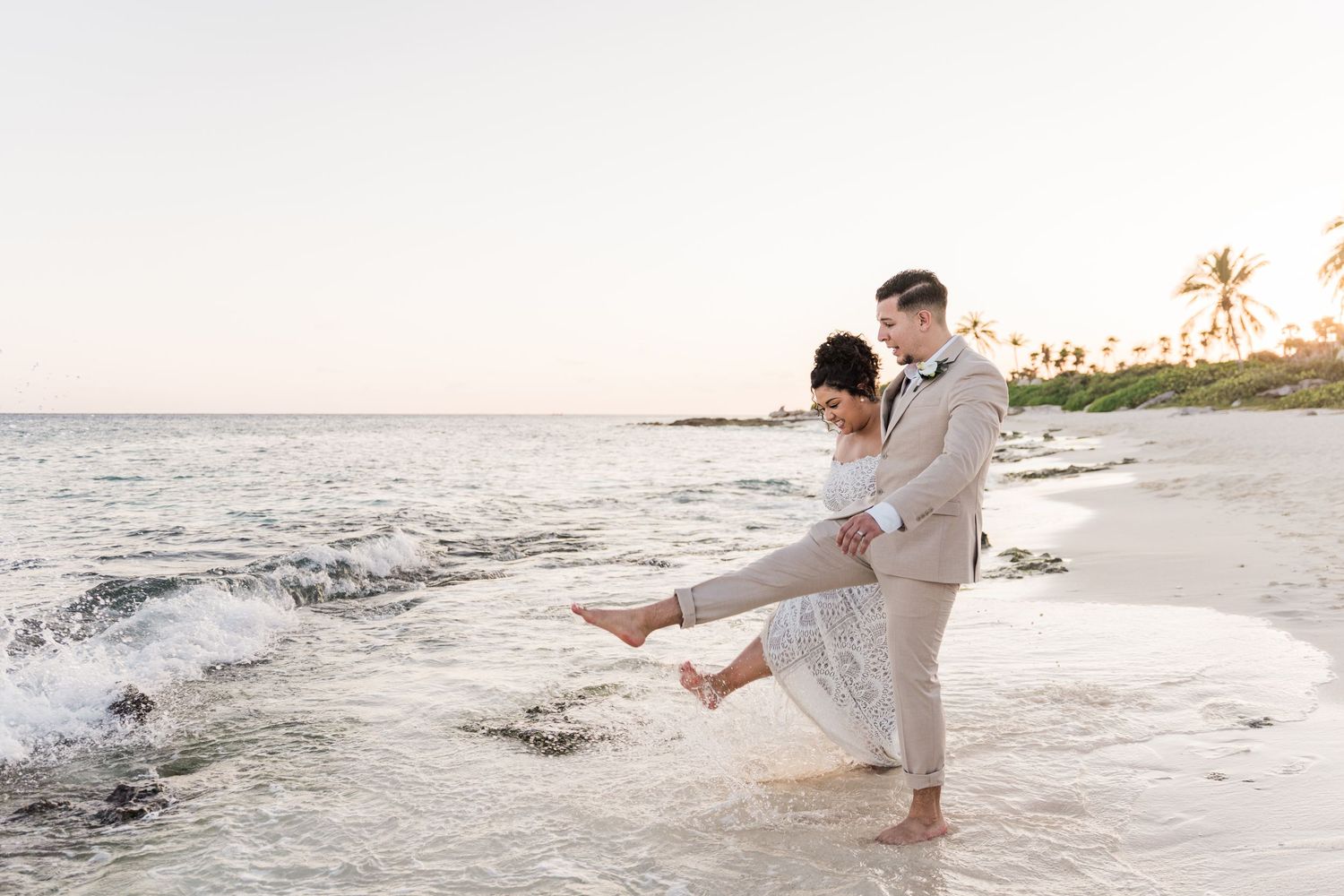 Xcaret Arte Wedding - Bride and groom laughing on a sandy beach at sunset, with ocean waves splashing around their bare feet in Hawaii.