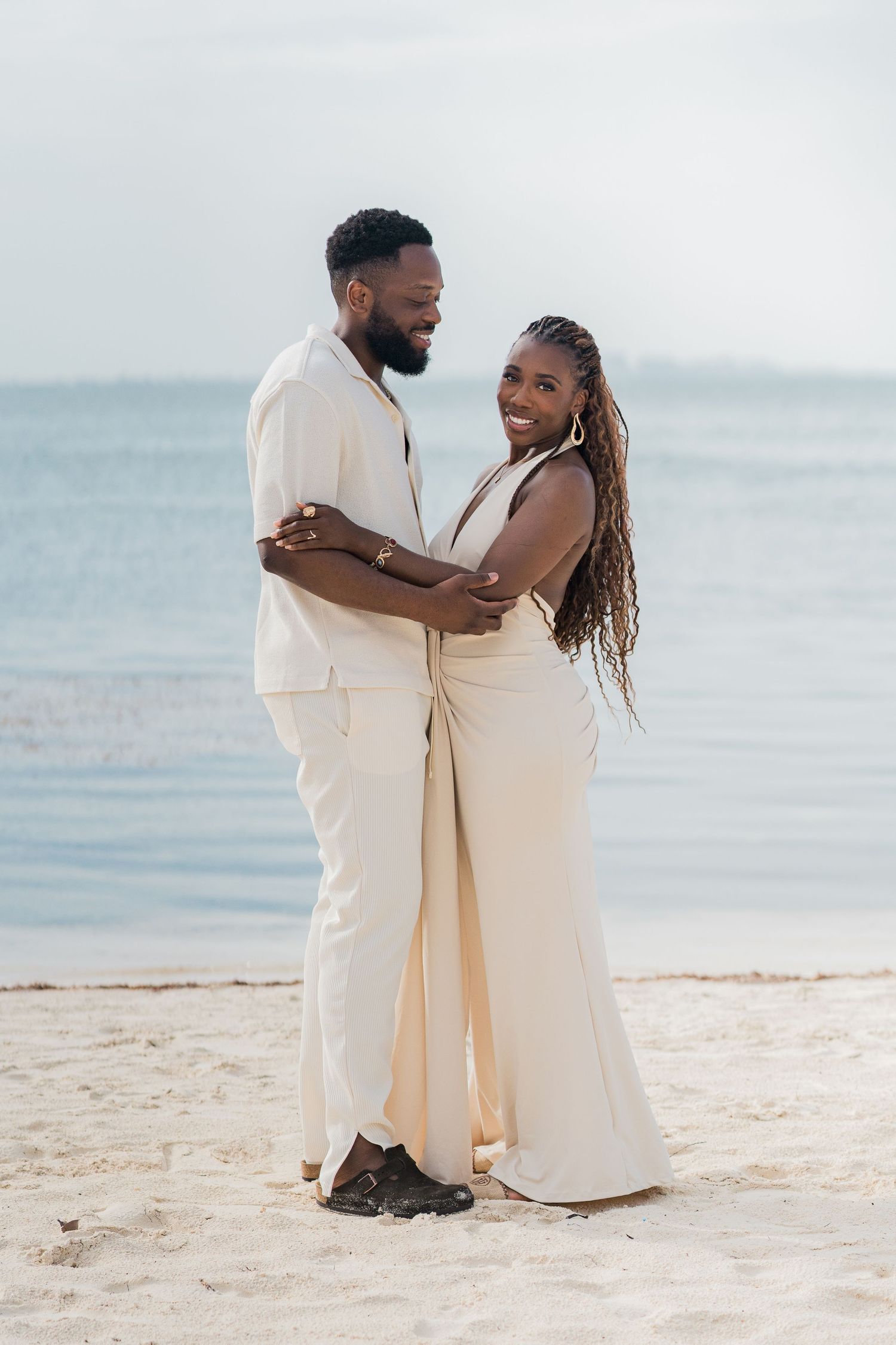 A couple in matching cream outfits embracing on a sandy beach with calm ocean waters in the background.