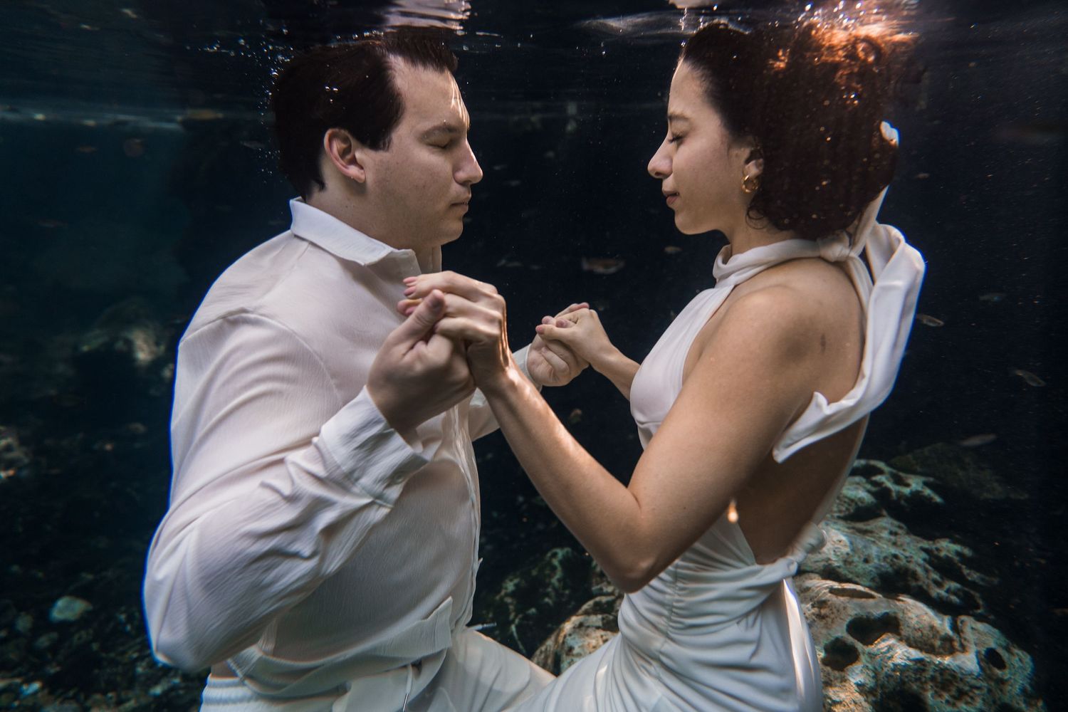 Couple in wedding attire holding hands underwater, surrounded by fish and coral in a romantic submerged photo shoot.