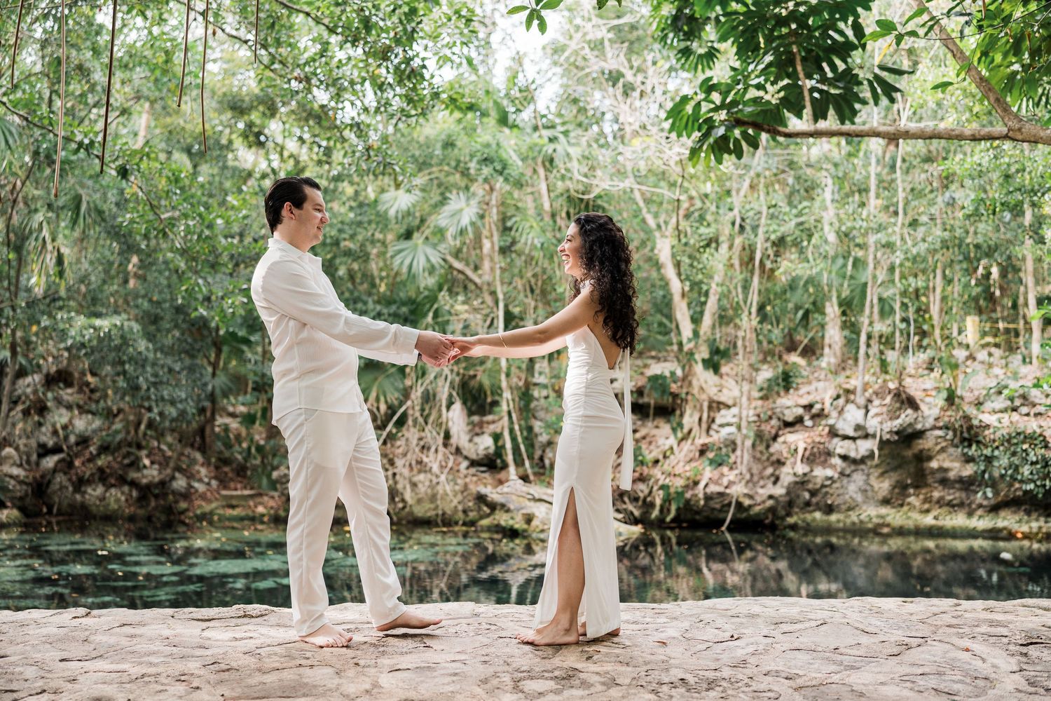 Couple in white outfits holding hands by a lush cenote surrounded by tropical trees.