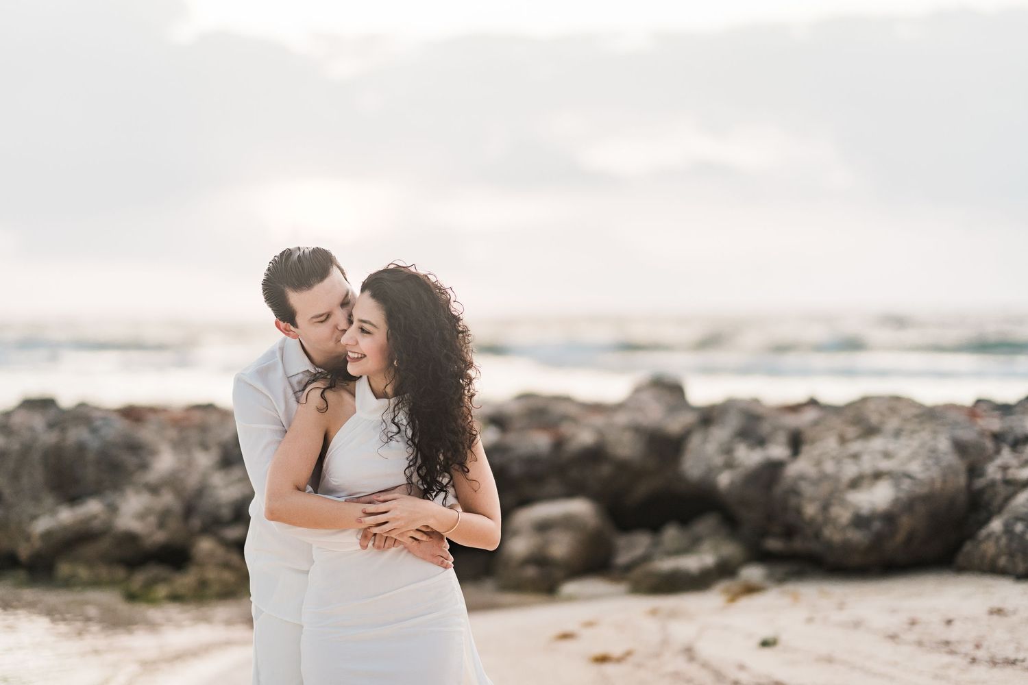 Couple embracing romantically on a sandy beach with rocky shoreline and ocean waves in the background.