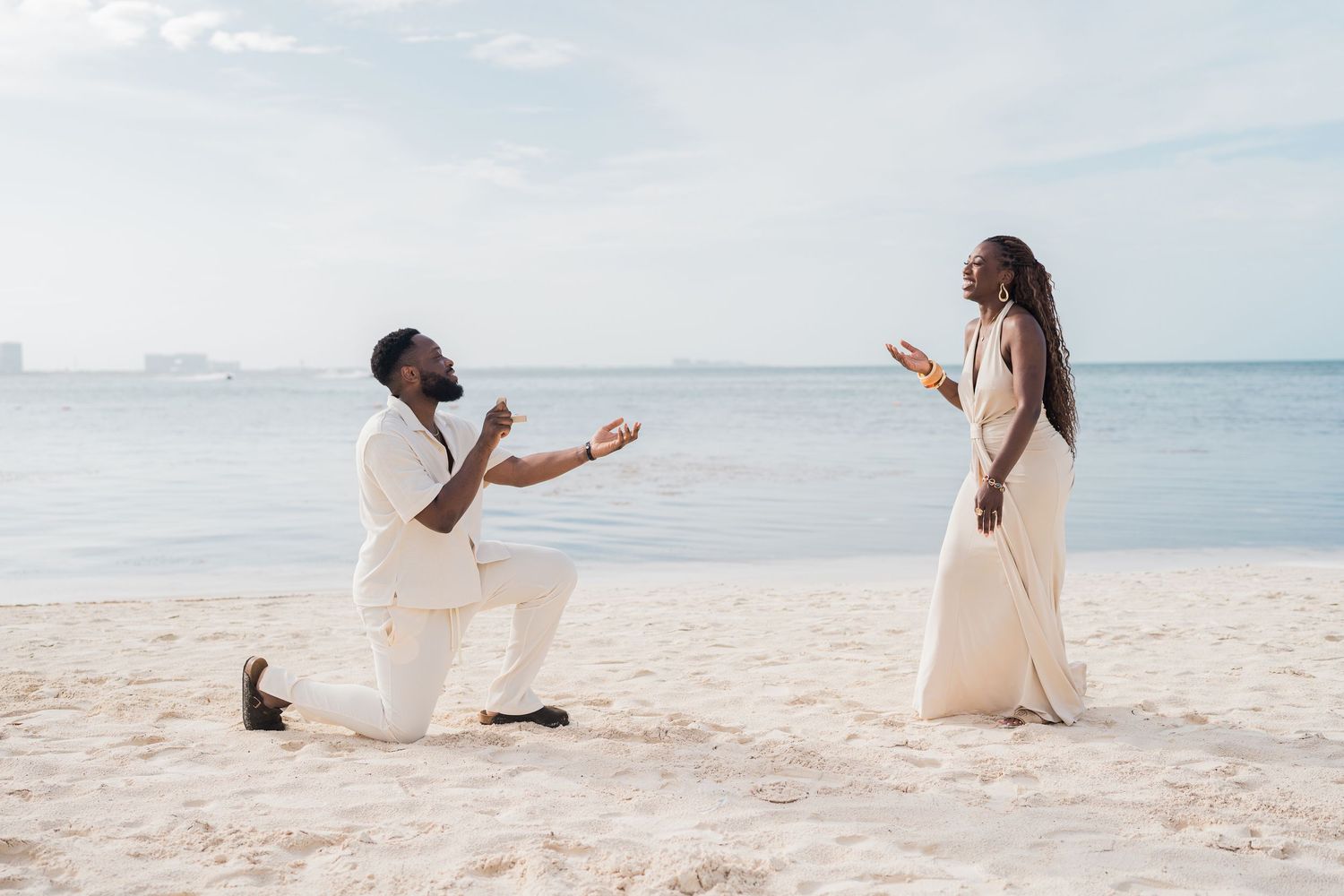 Man proposing on one knee to woman in white dress on sandy beach with ocean backdrop.