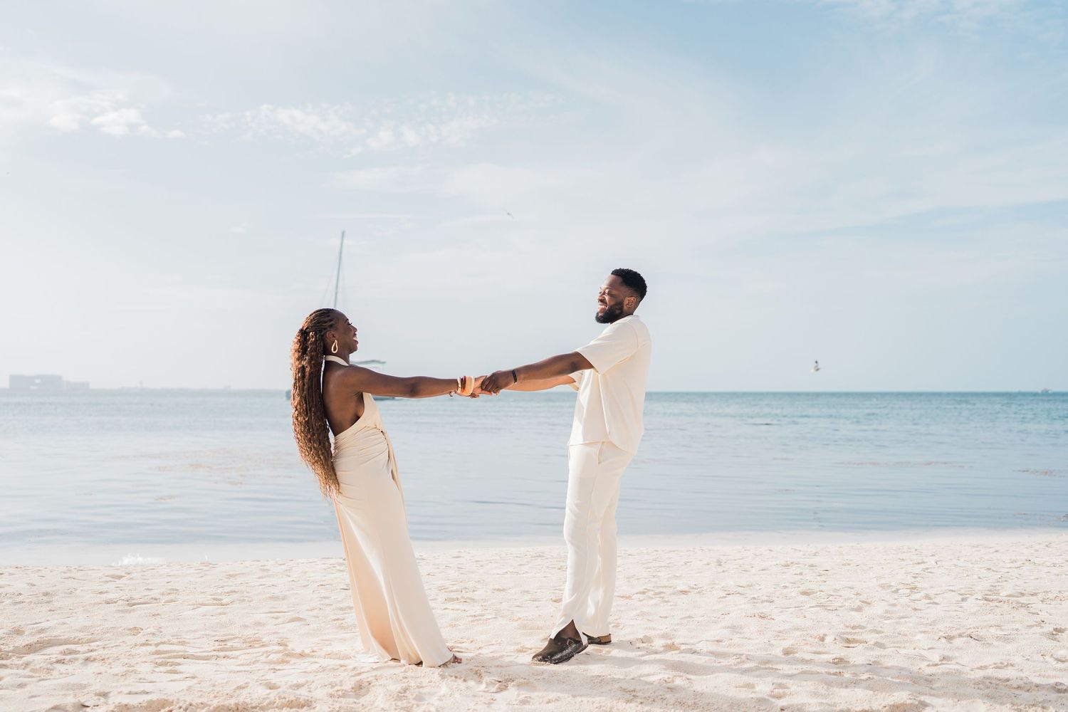 Couple in white outfits holding hands on a sunny beach with calm ocean waters in the background.