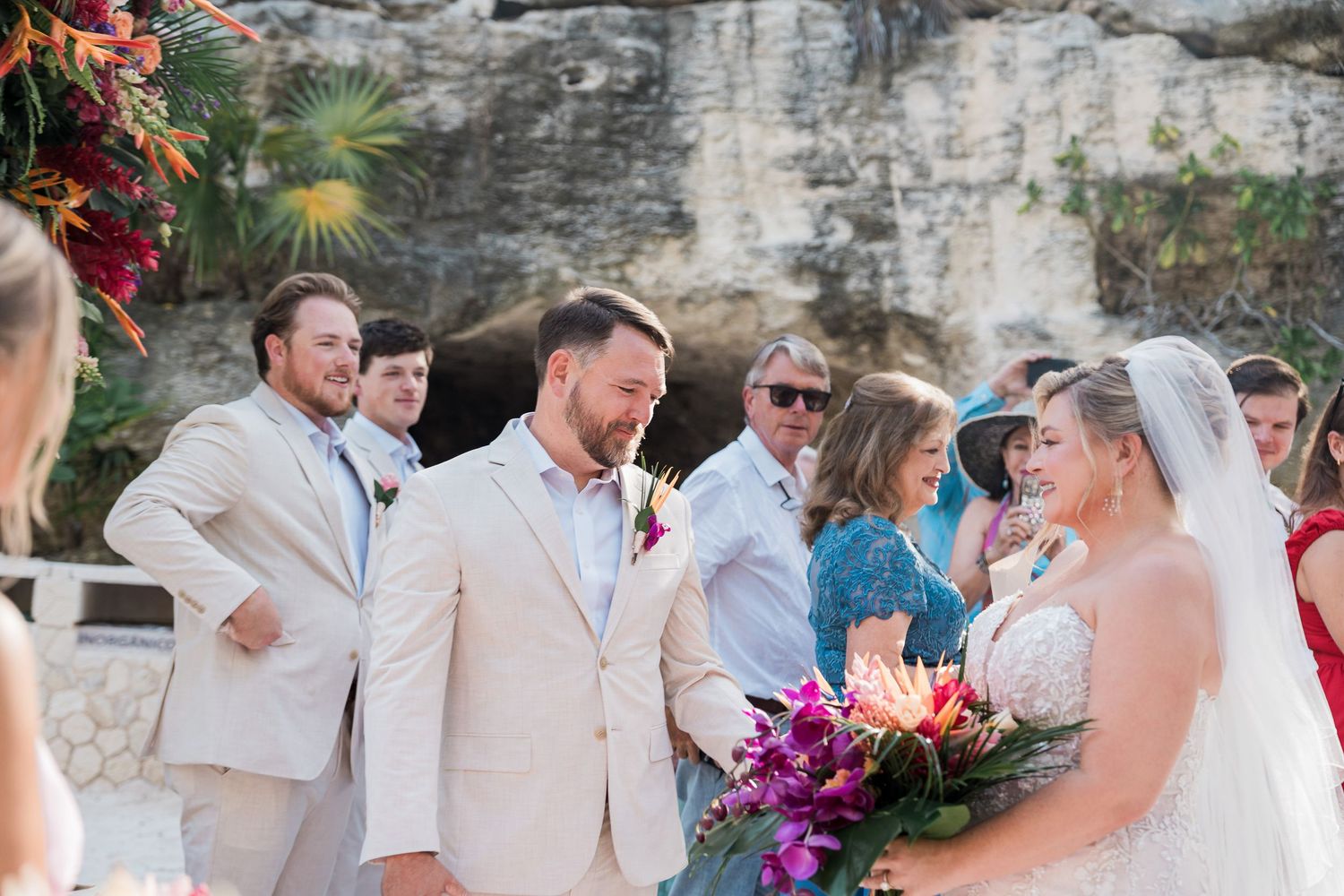Groom in beige suit smiles at bride holding tropical bouquet during outdoor wedding ceremony with guests.
