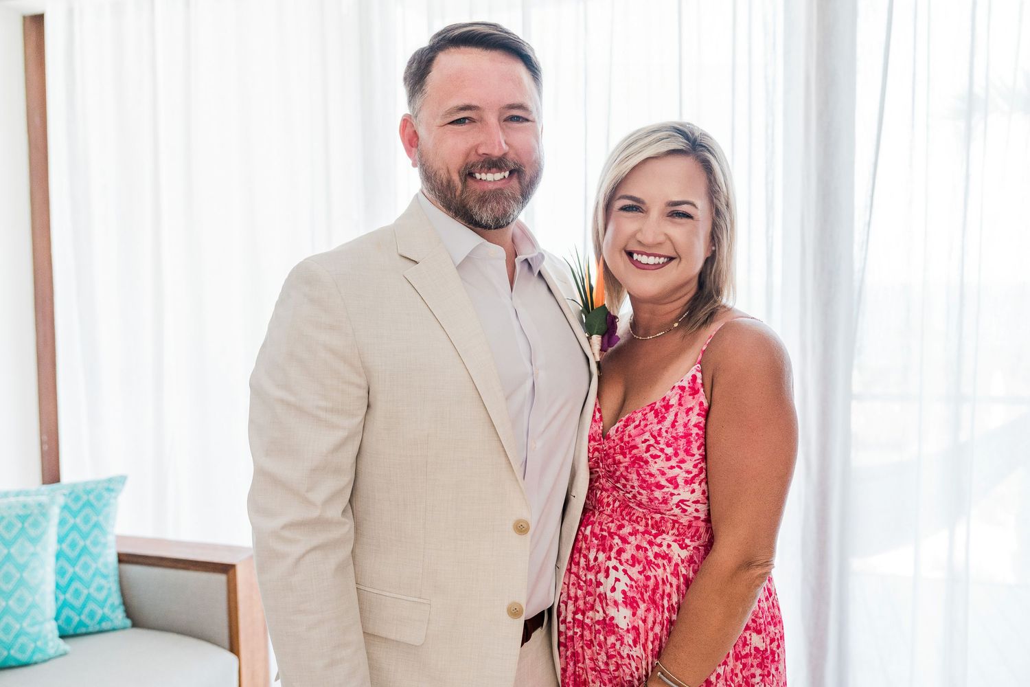 Smiling couple posing together, man in beige suit and woman in pink floral dress, bright modern interior backdrop.