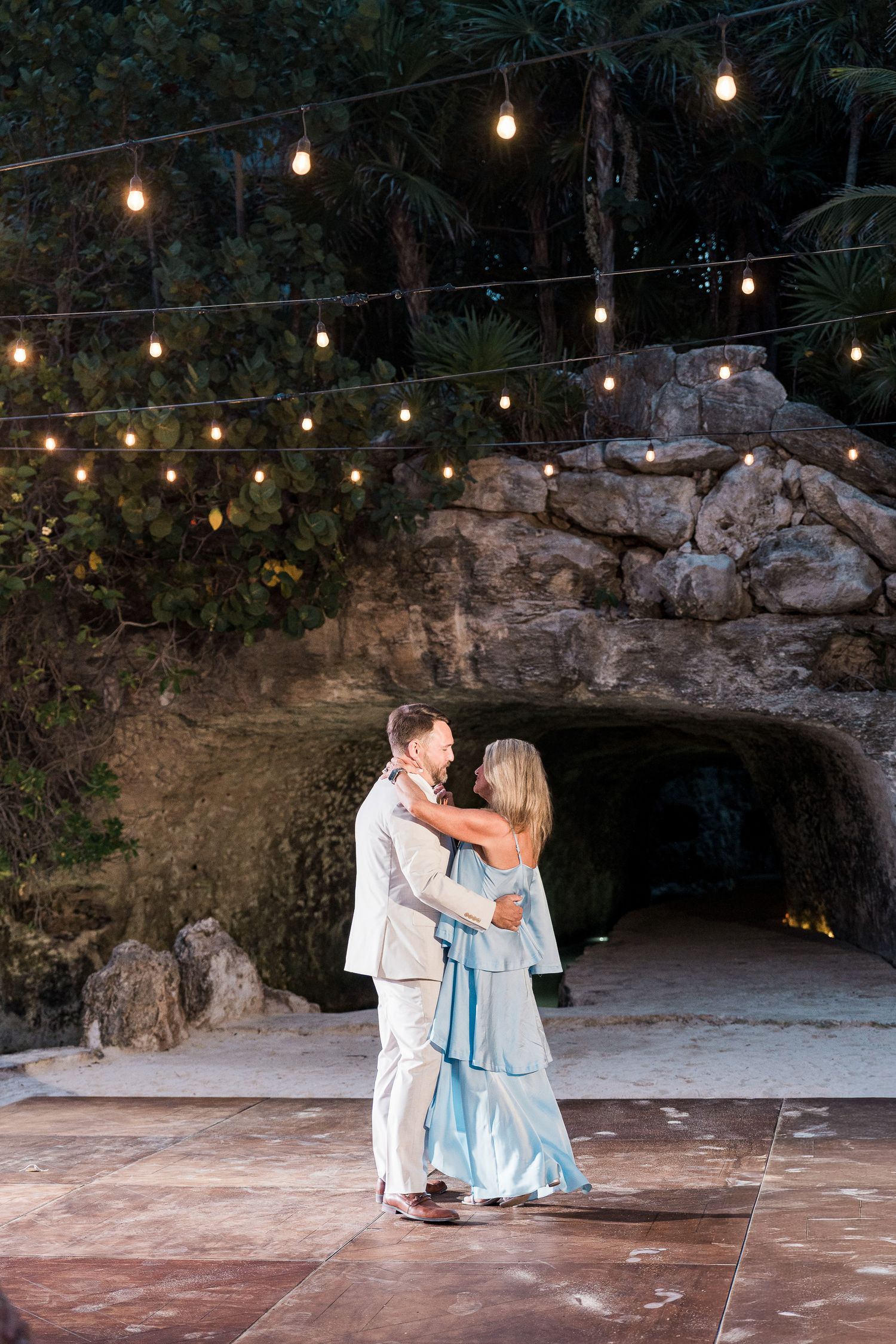 Couple dancing outdoors at night under string lights near a stone grotto at a romantic evening celebration.
