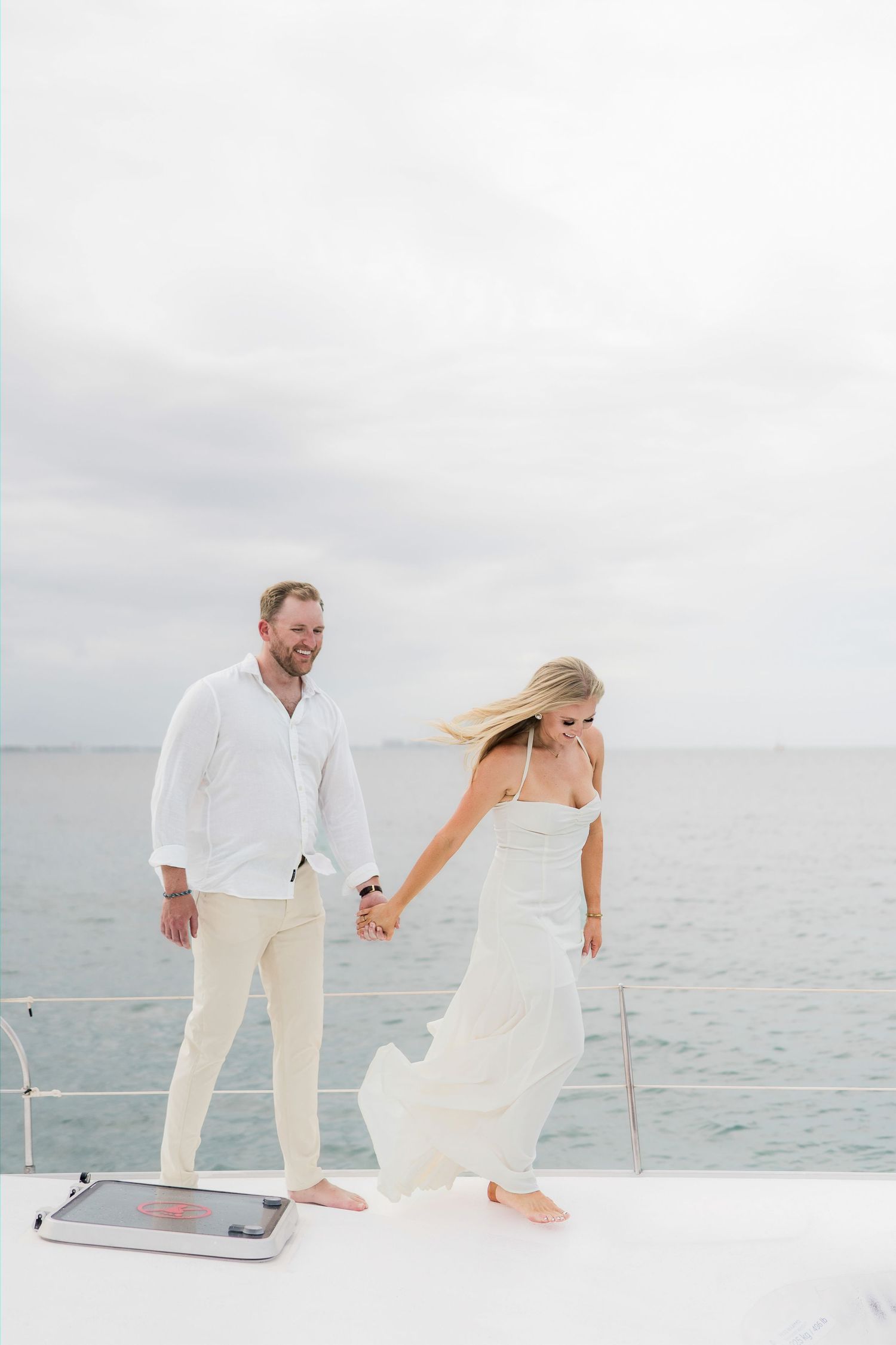 Cancun Engagement Photoshoot - Couple in white outfits holding hands on a yacht deck with ocean backdrop on a cloudy day.