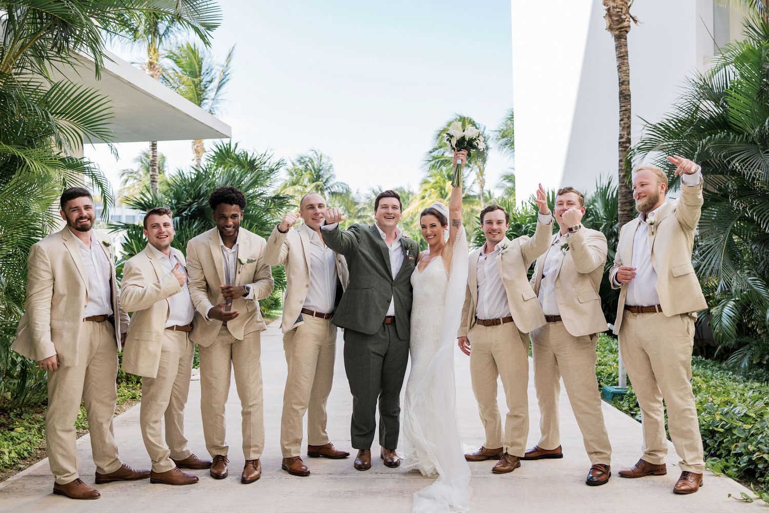 Bride and groom pose with groomsmen in tan suits outdoors at a tropical wedding venue with palm trees.