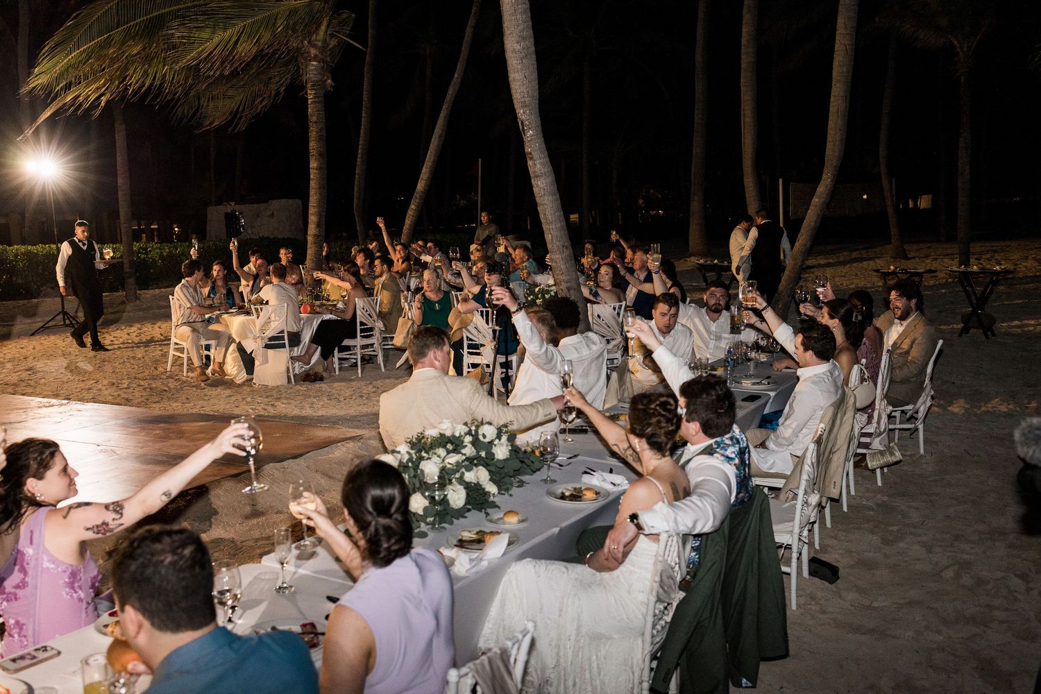 Guests raising glasses in a toast at an outdoor beach wedding reception at night, surrounded by palm trees and elegant table settings.