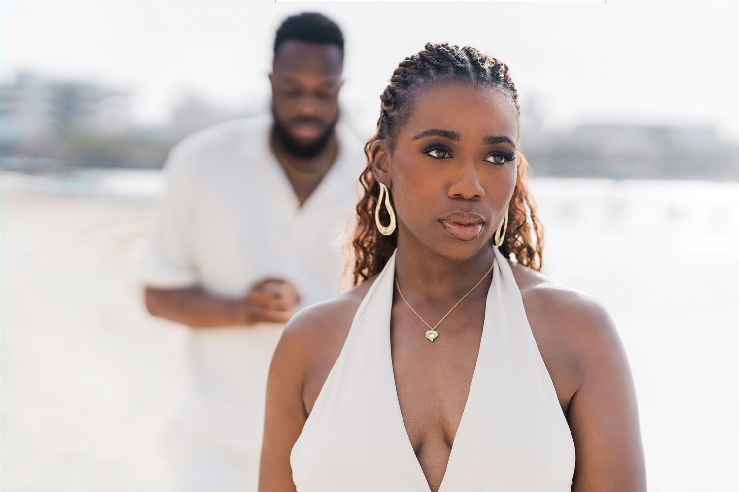 A woman in a white halter dress stands in the foreground, with a man in white attire blurred in the background.