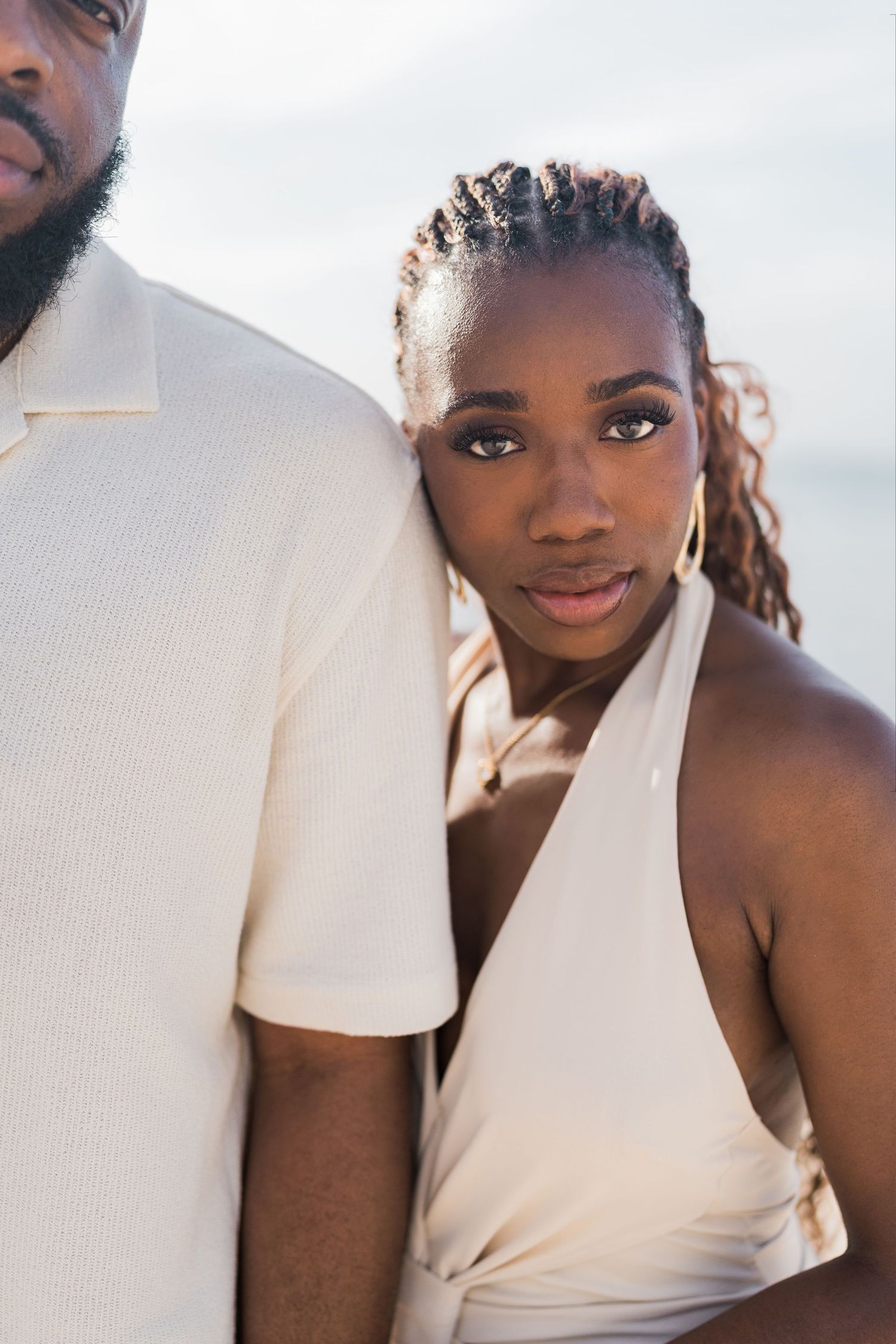 A Black couple in cream outfits pose closely together on a beach, the woman leaning against the man's shoulder.