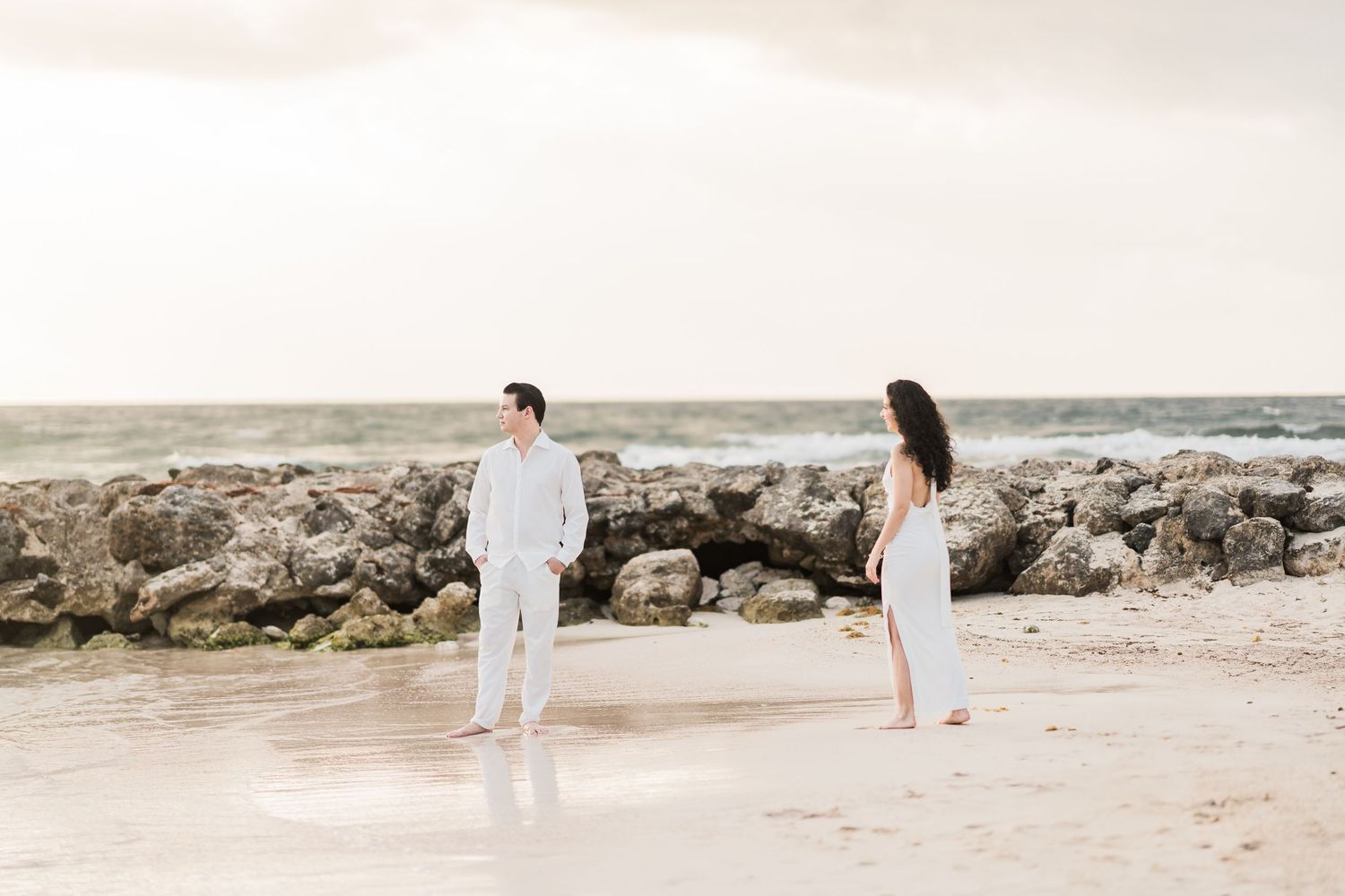 A couple in white outfits stands on a sandy beach near rocky shores with ocean waves in the background.