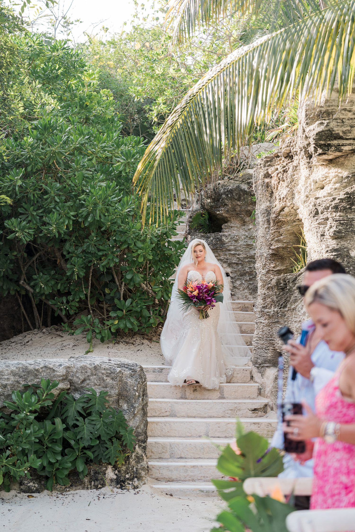 Hotel Xcaret México Wedding  -Bride in a white gown descends stone steps holding a colorful bouquet at an outdoor tropical wedding ceremony.