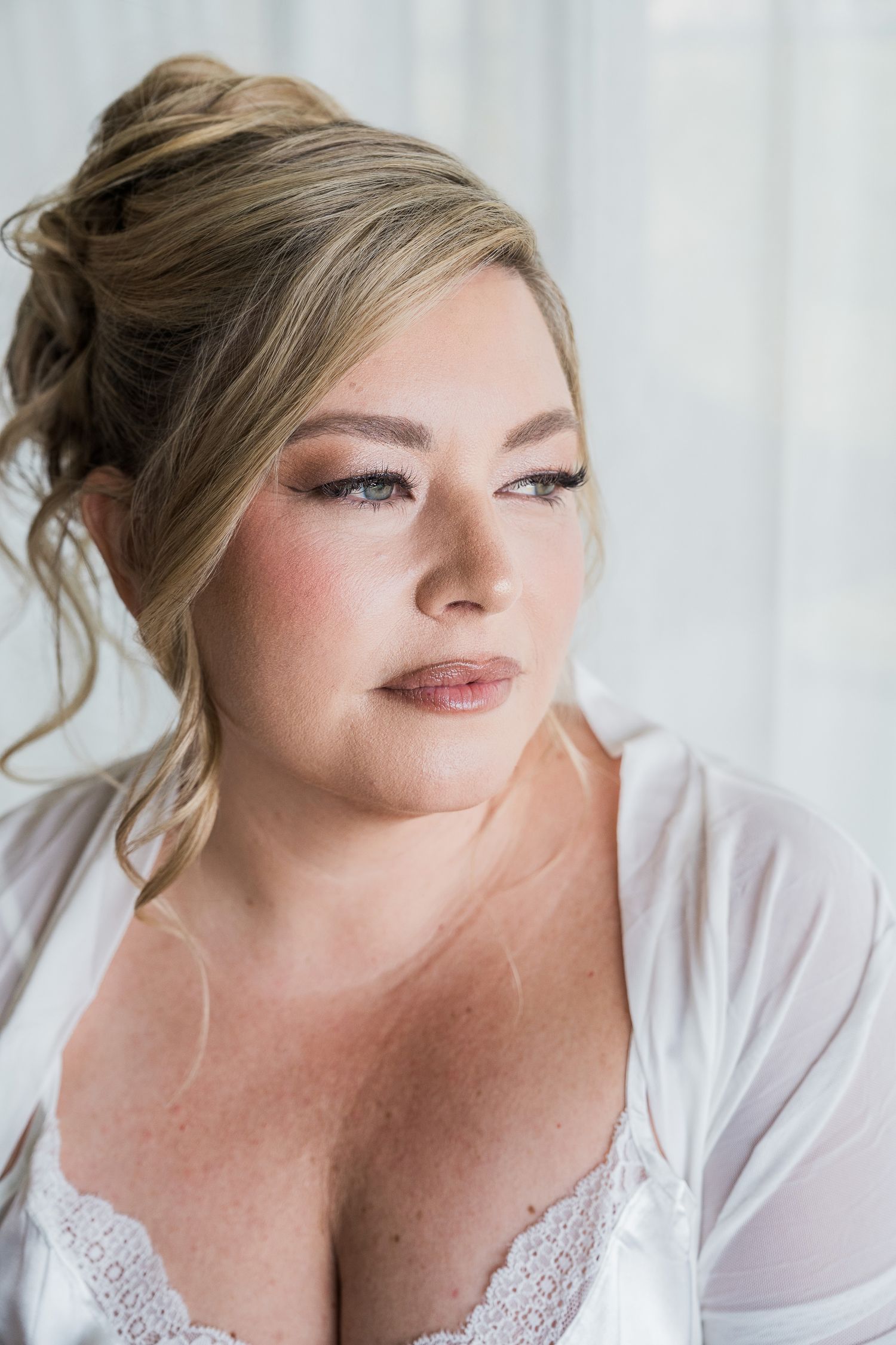 Hotel Xcaret México Wedding.Blonde woman in white lace bridal robe with elegant updo and soft glam makeup gazing thoughtfully near a window.