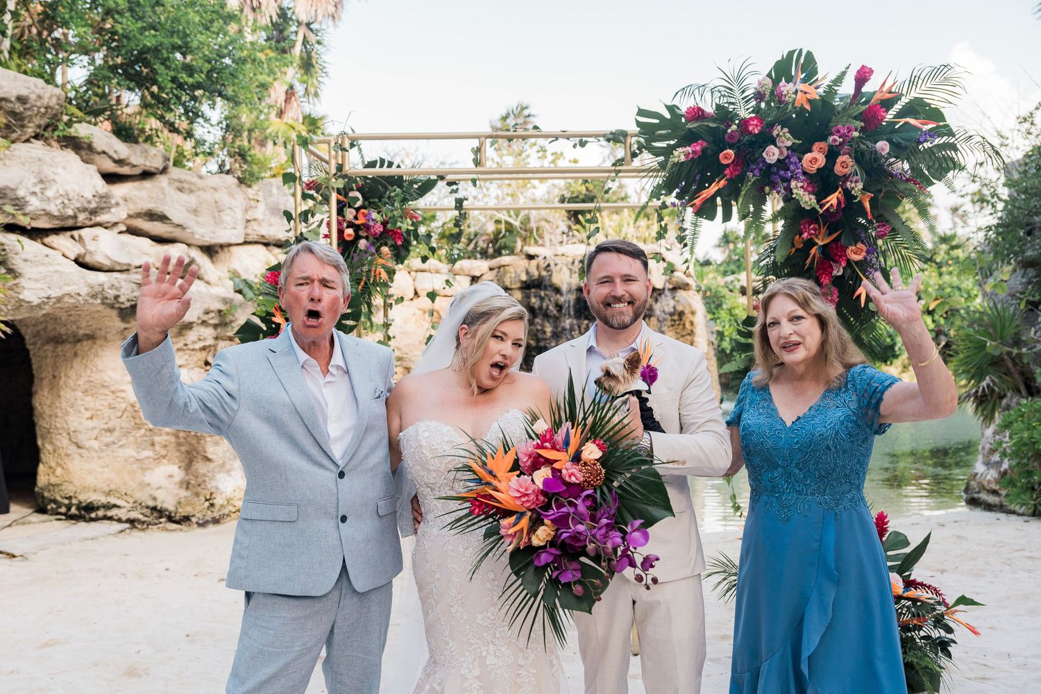Joyful wedding party with bride holding tropical bouquet celebrating in front of floral arch at outdoor venue.