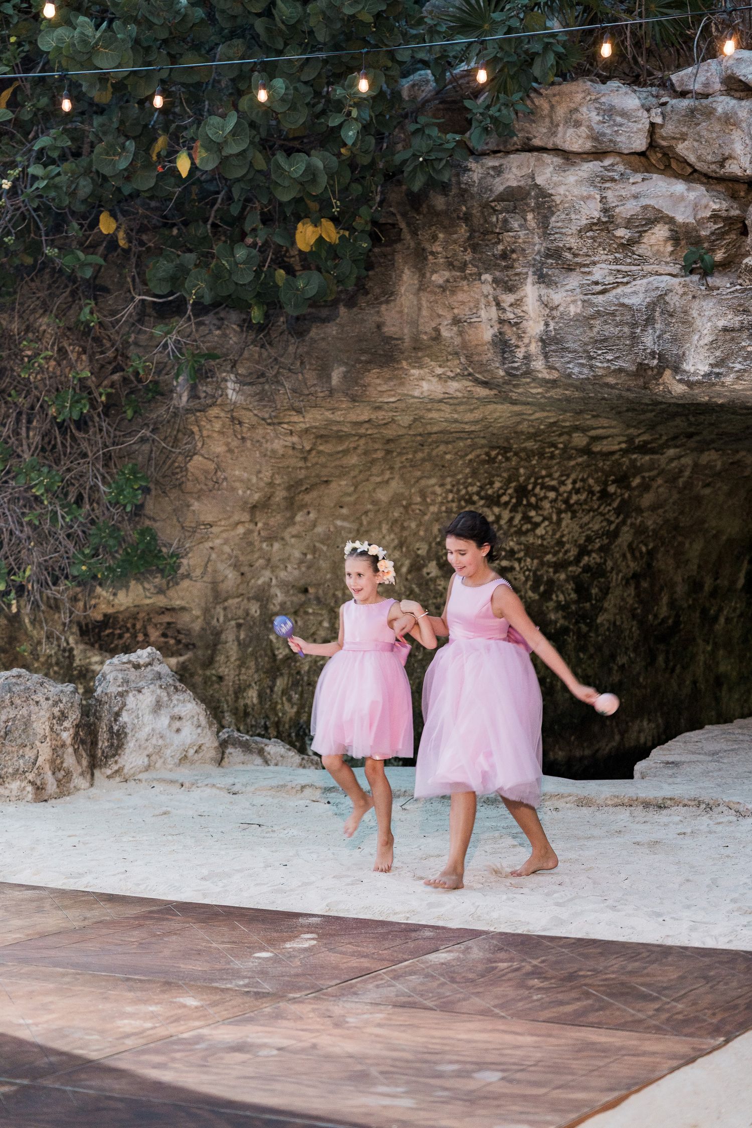 Two flower girls in pink tulle dresses dance barefoot on a sandy beach near a rocky cave backdrop with string lights.