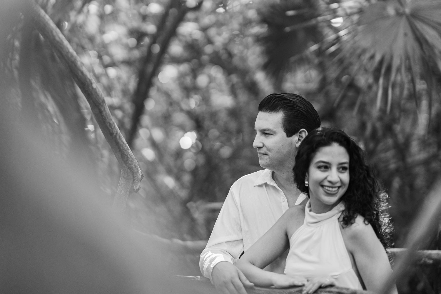 Black and white photo of a smiling couple embracing outdoors among tropical palm trees and lush greenery.