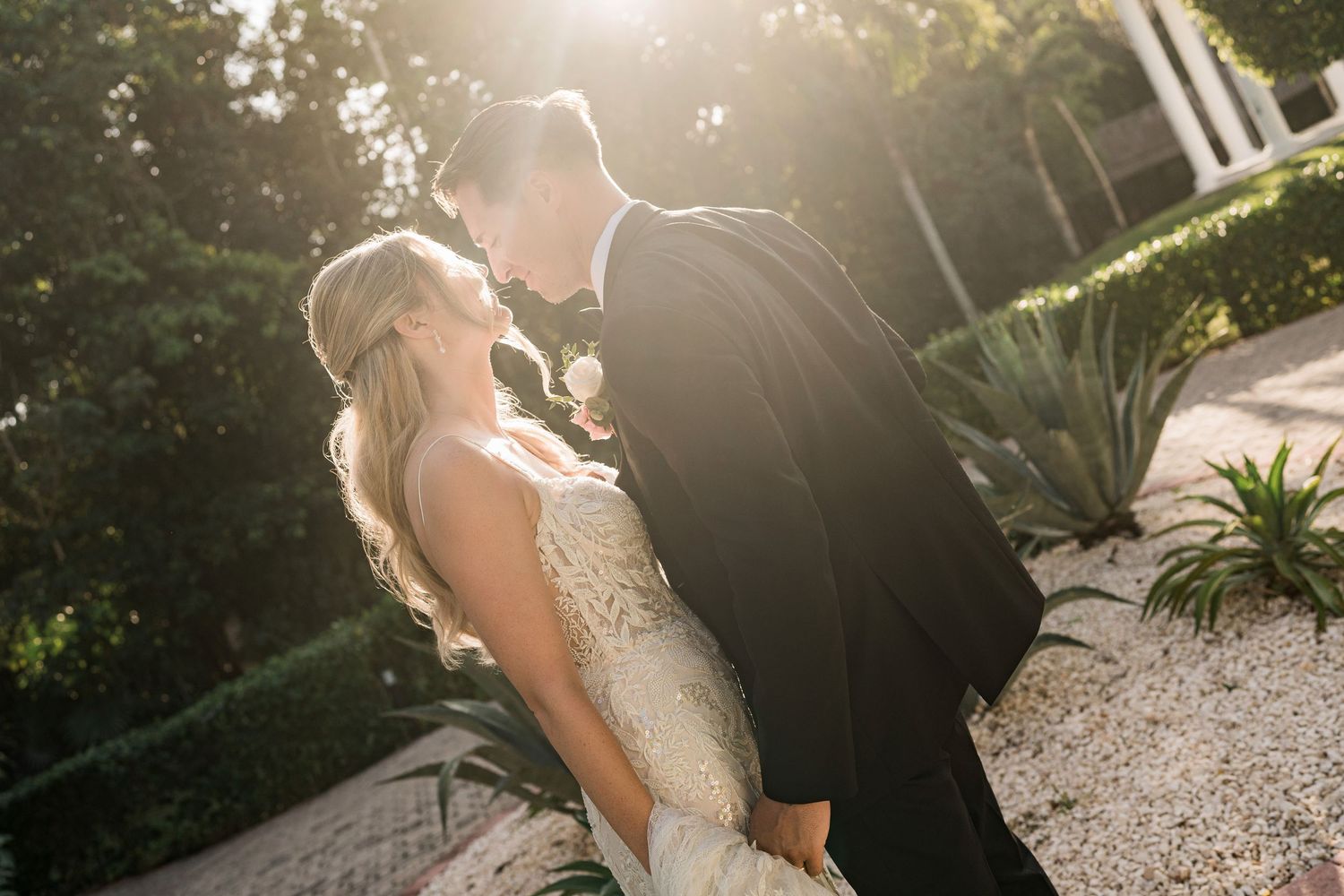 Bride and groom share a romantic moment in golden sunlight, surrounded by lush greenery at an outdoor wedding venue.