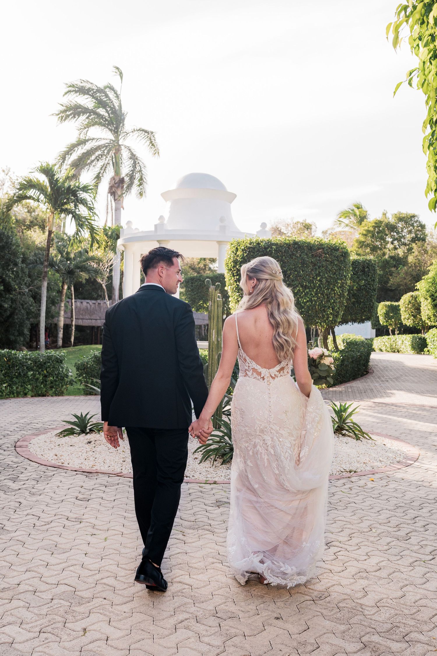 Bride and groom walking hand in hand toward a white gazebo surrounded by palm trees at a tropical wedding venue.