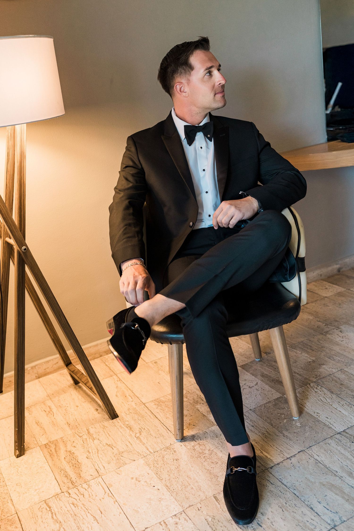 A man in a black tuxedo with bow tie sits elegantly on a chair, holding accessories, gazing thoughtfully to the side.
