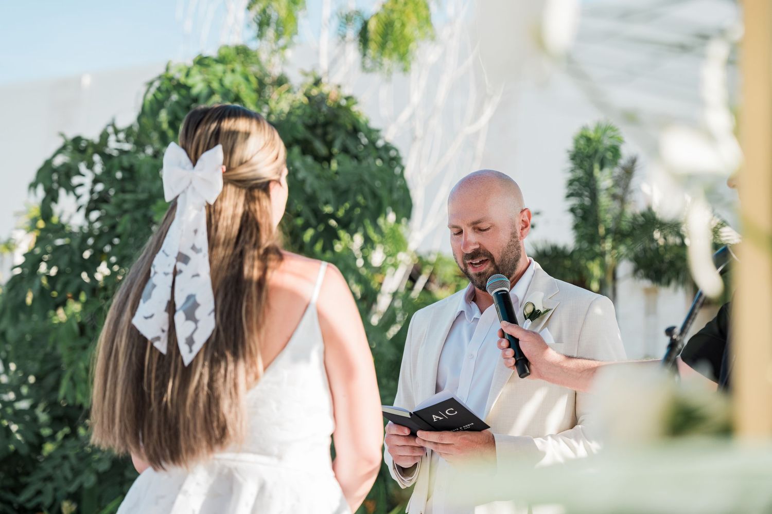 Groom in white suit reads vows to bride with bow in hair at outdoor wedding ceremony with greenery backdrop.