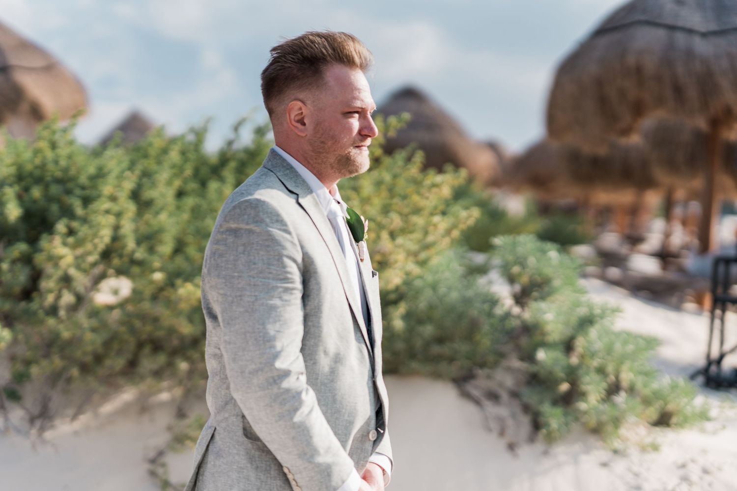 Groom in gray suit standing on sandy beach with tropical thatched huts and greenery in background.