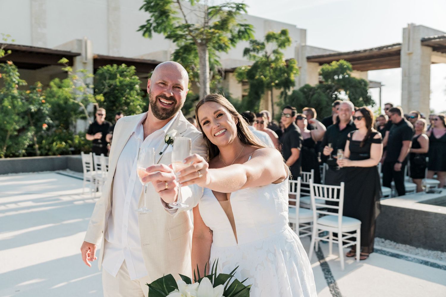 Bride and groom smiling and toasting with drinks at outdoor wedding ceremony on sunny rooftop venue.