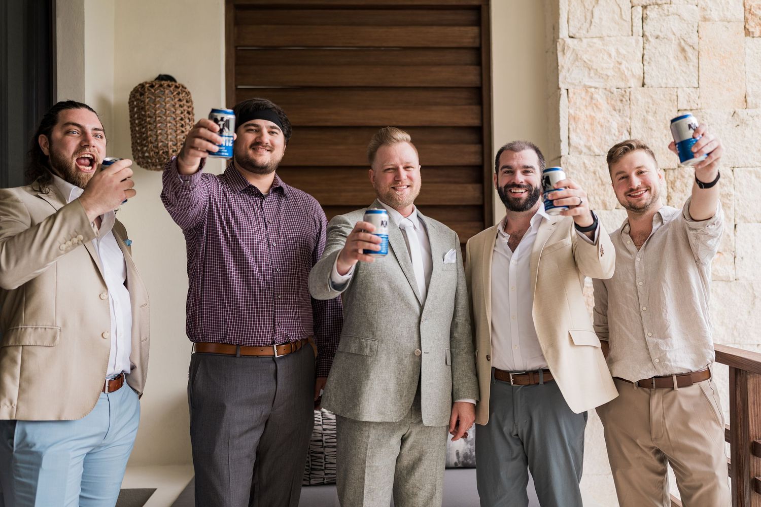 Five men smiling and raising canned drinks in a toast, dressed in smart casual attire at an outdoor venue.