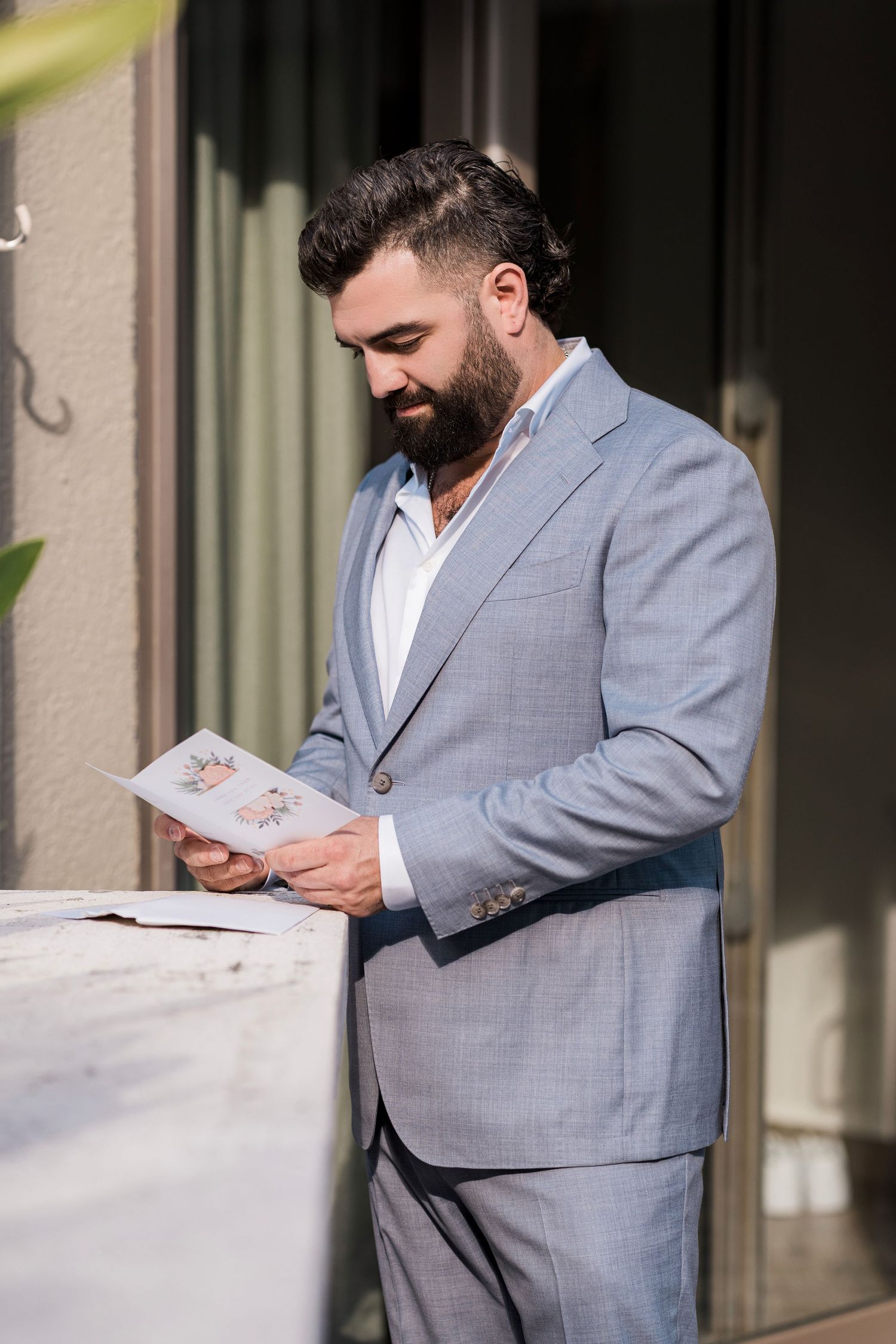 Bearded man in light gray suit reading a menu card outdoors at a restaurant terrace in bright sunlight.