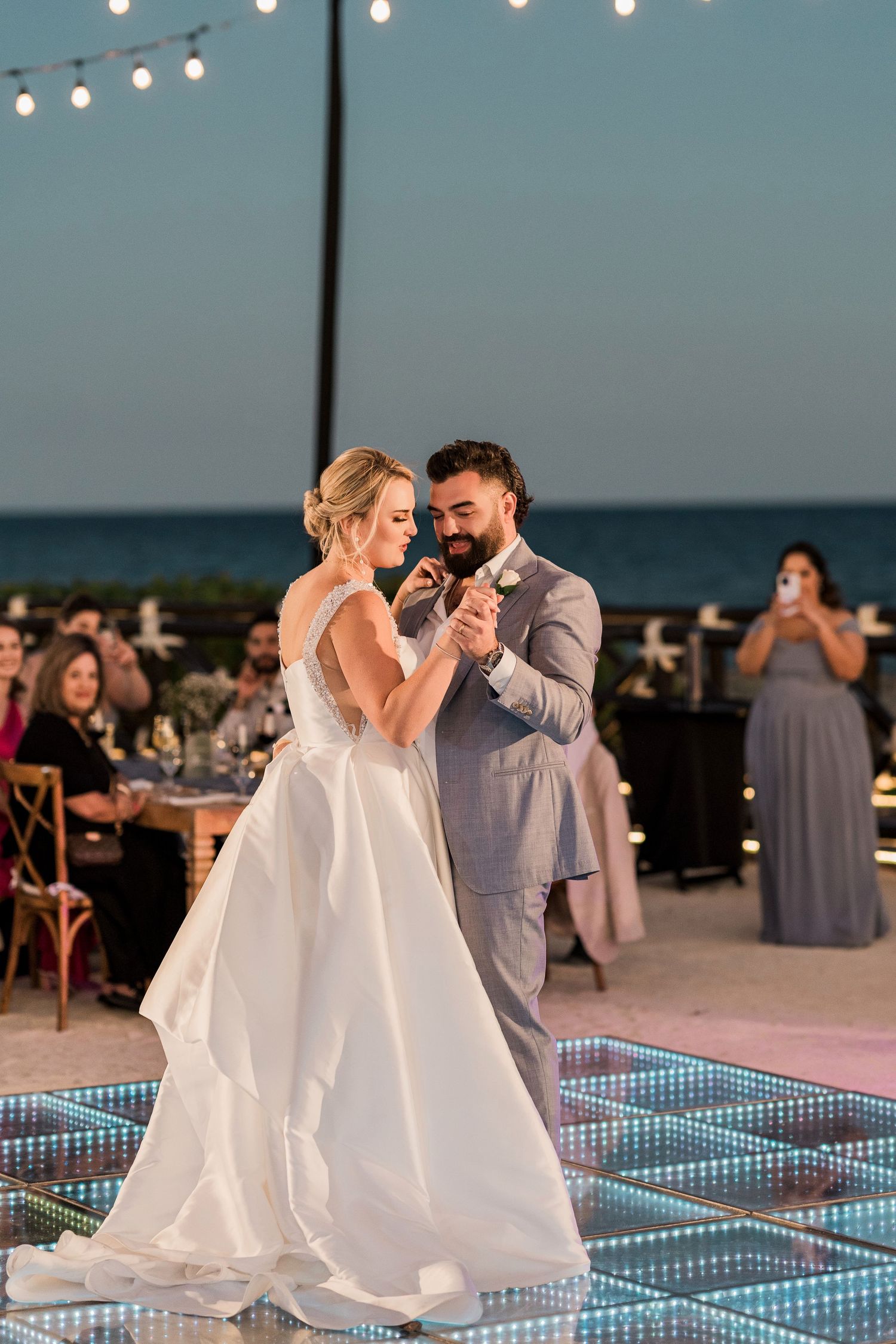 Bride and groom share their first dance on an illuminated floor at an outdoor beach wedding reception at dusk.