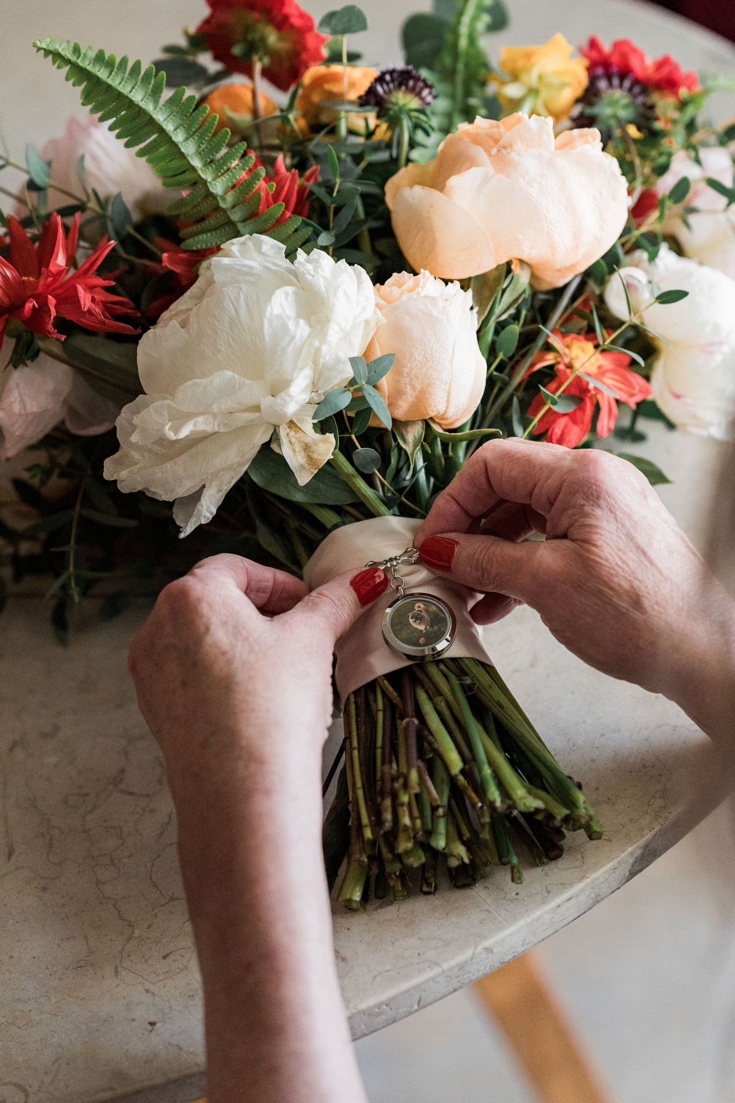 Florist tying a ribbon around a colorful wedding bouquet with peonies and tropical flowers on a marble table.