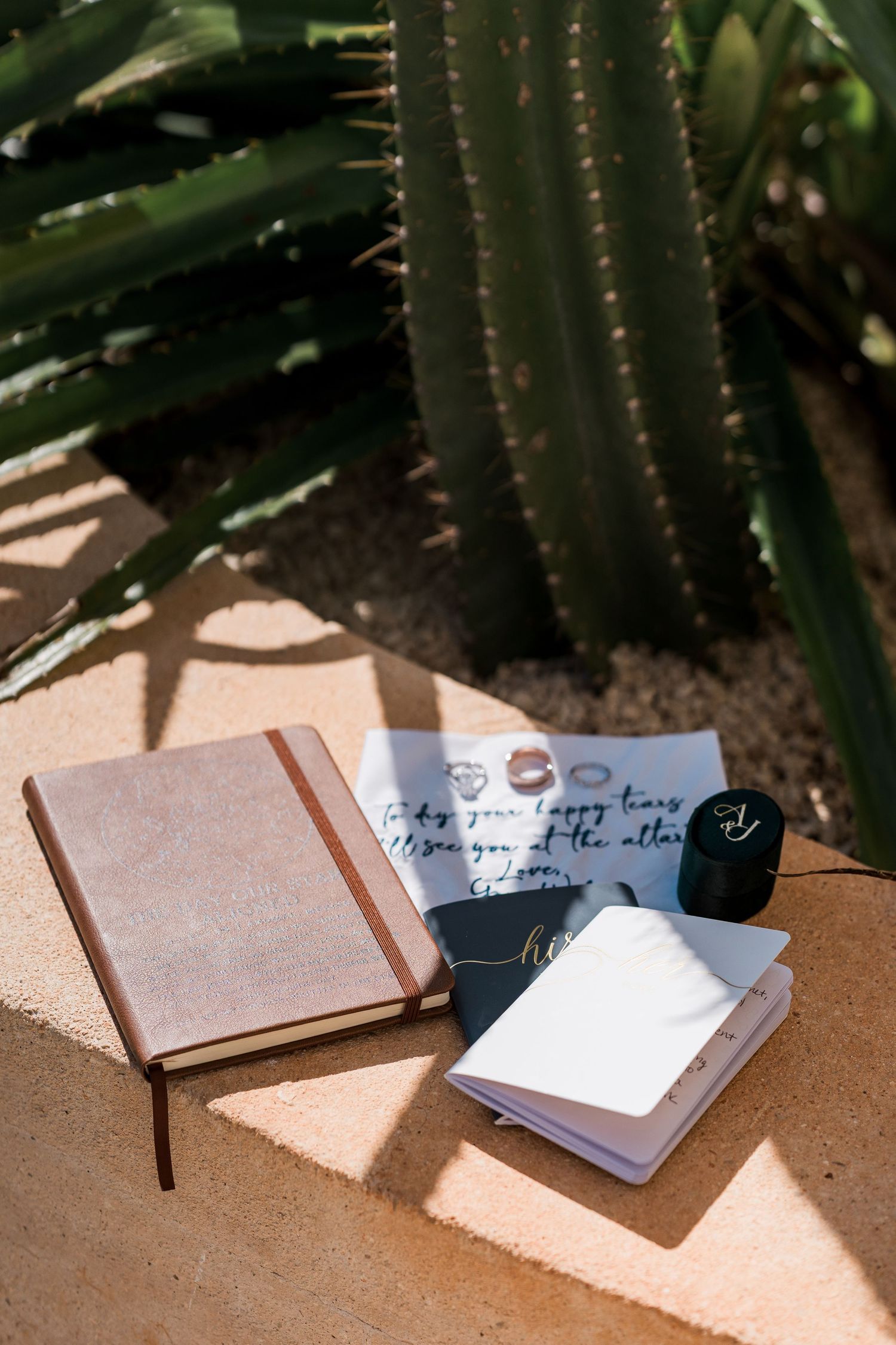 Secrets Akumal Wedding - Leather journal, open notebook, and handwritten note arranged on stone surface beside a large cactus in sunlight.