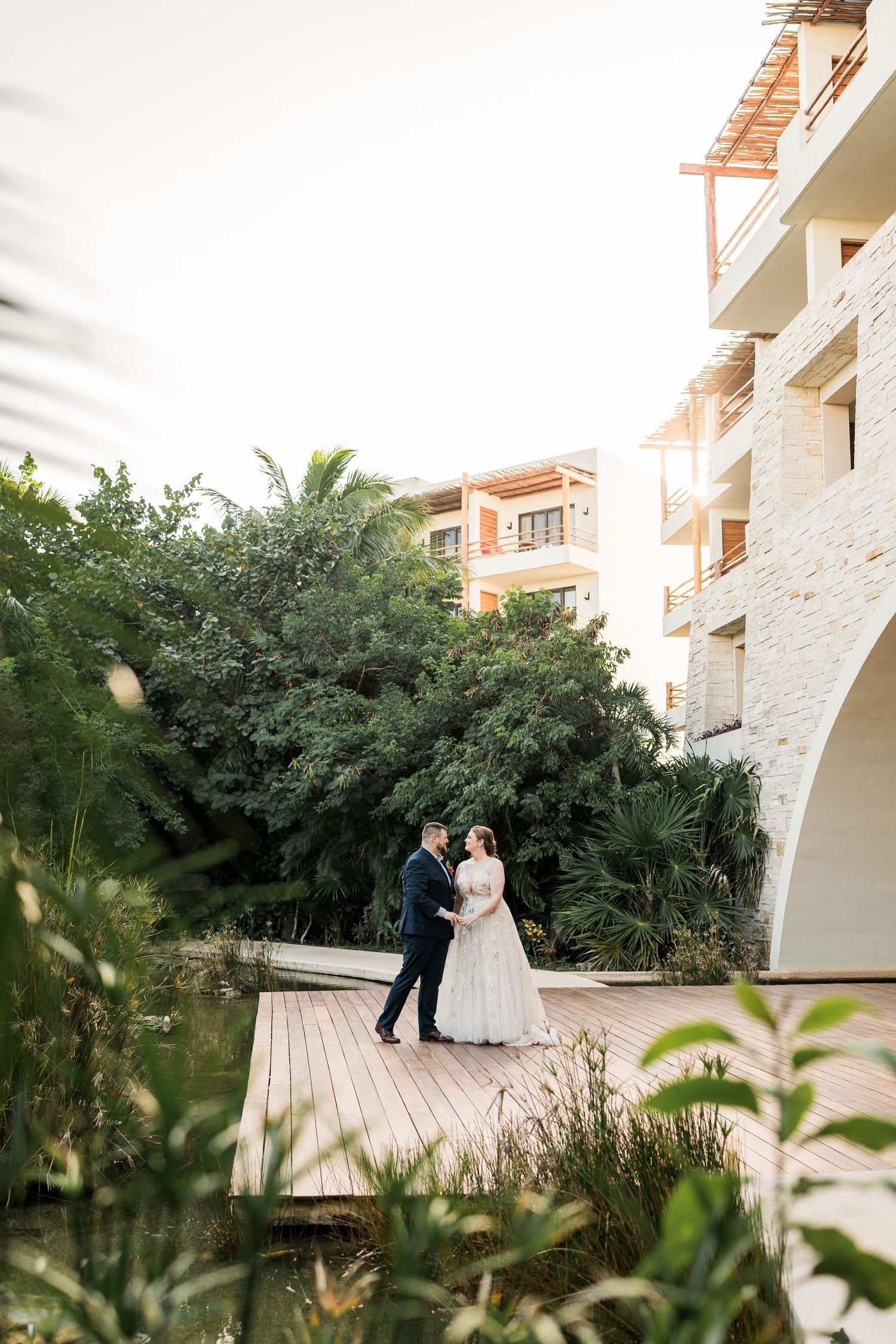 Secrets Akumal Wedding - Bride and groom embracing on a wooden deck surrounded by lush greenery and a luxury resort building.