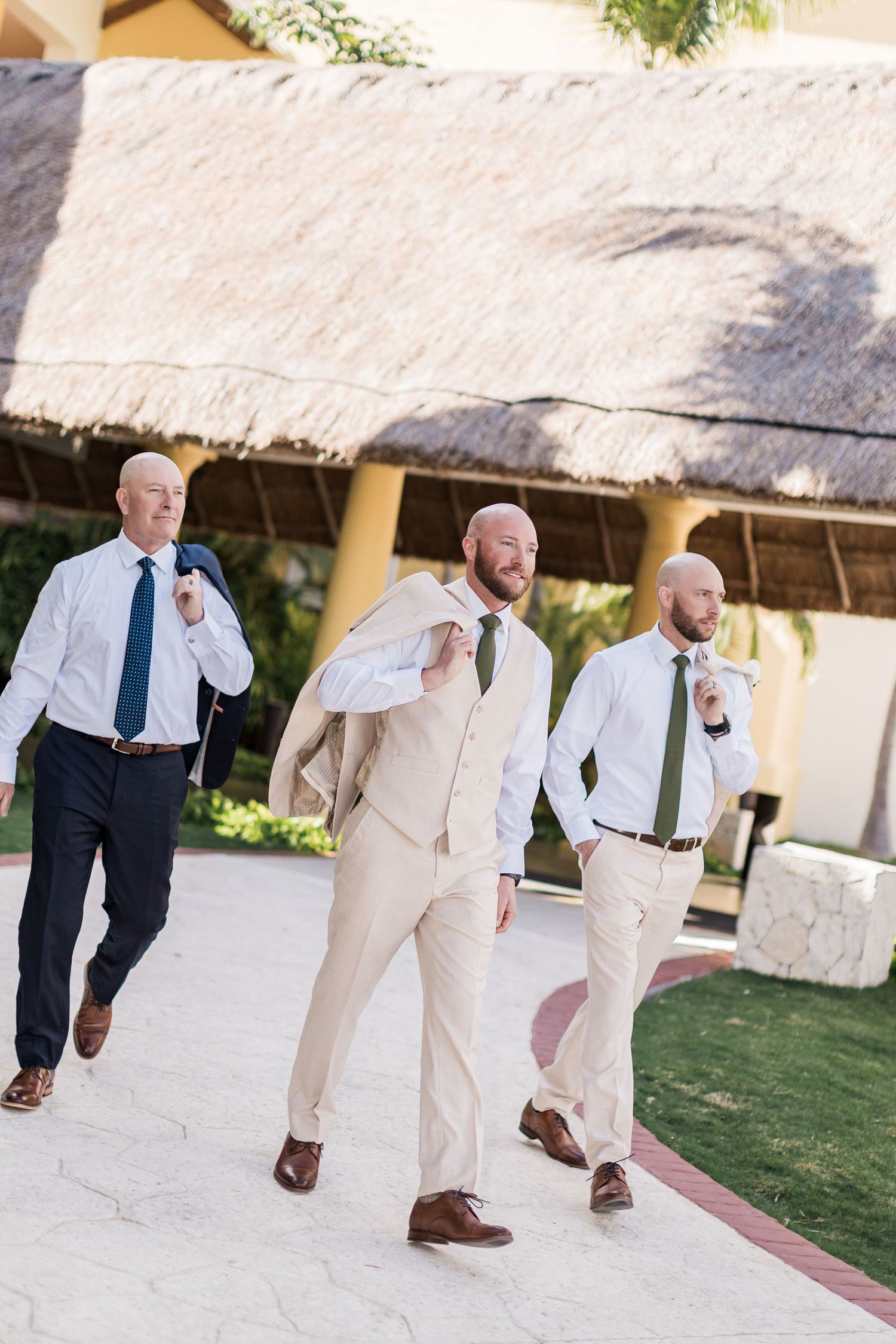 Three bald groomsmen in tan and navy suits with green ties walk confidently at an outdoor tropical wedding venue.