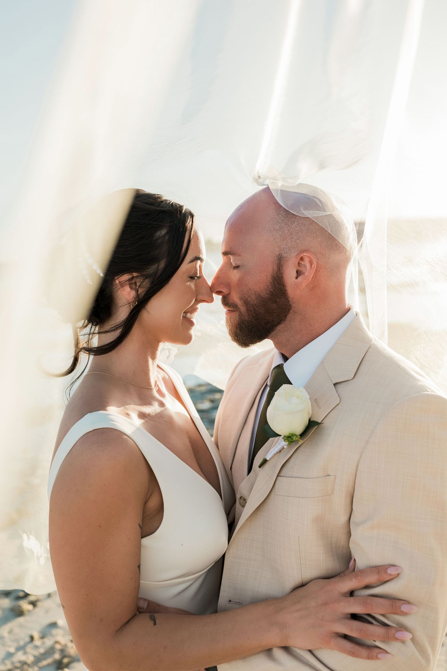 A bride and groom share a romantic moment under a flowing veil at a beach wedding, with golden sunlight behind them.
