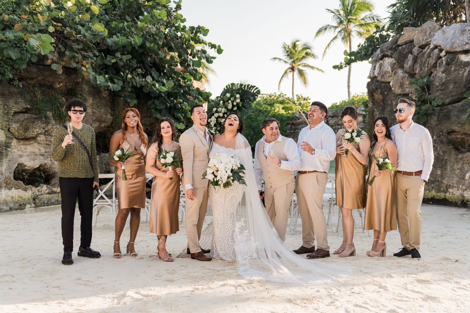 Xcaret Arte Wedding - Wedding party poses on sandy beach in gold bridesmaid dresses and neutral suits with tropical greenery backdrop.