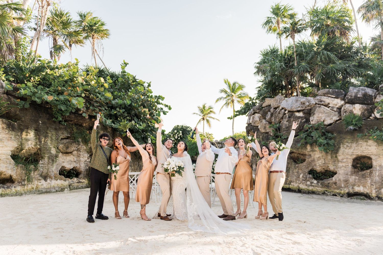 Xcaret Arte Wedding - Wedding party celebrating on a tropical beach with palm trees, bride in white gown with veil, guests in neutral tones.