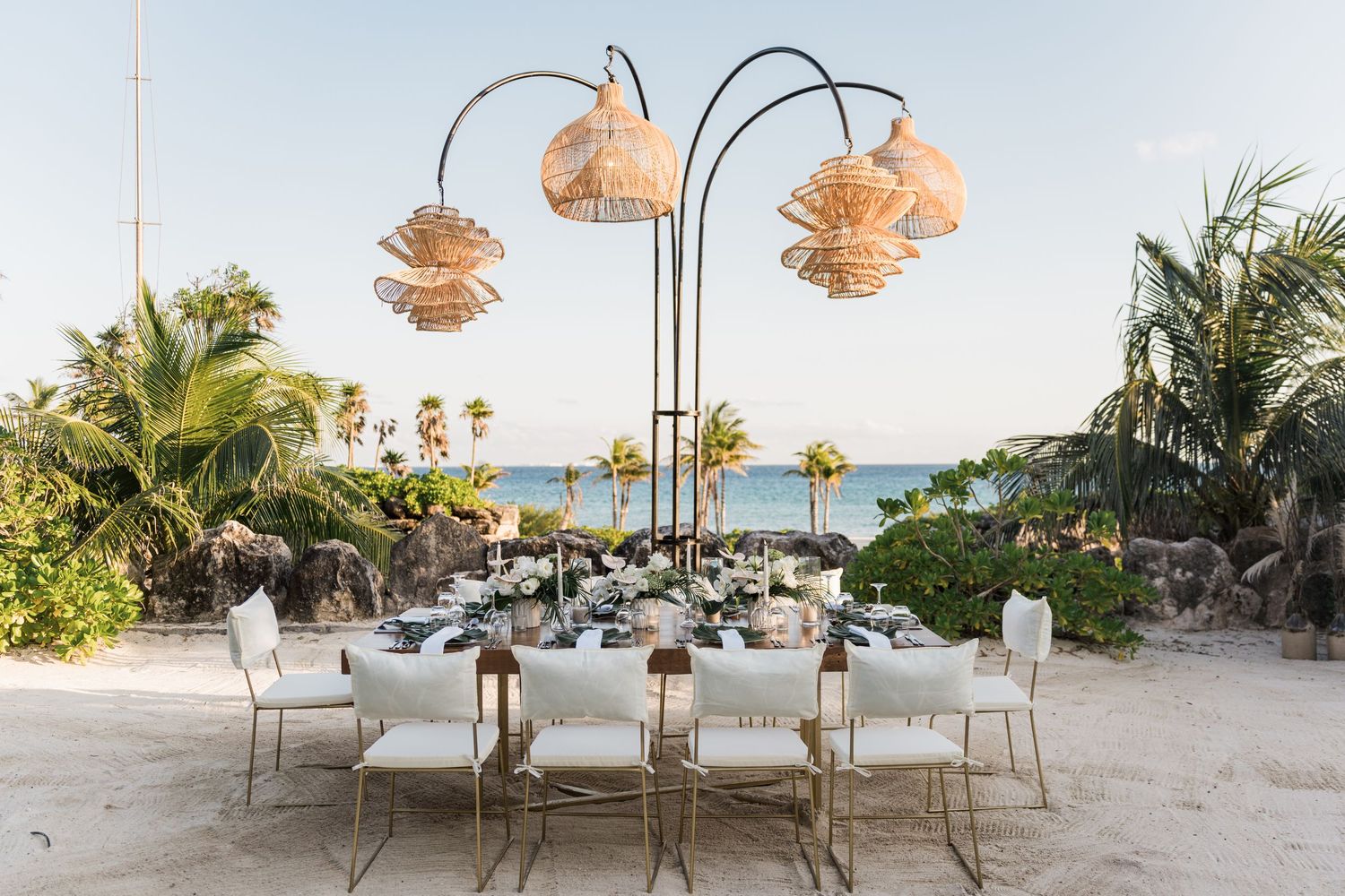 Xcaret Arte Wedding - Elegant beachside dining table set under woven pendant lights with white chairs